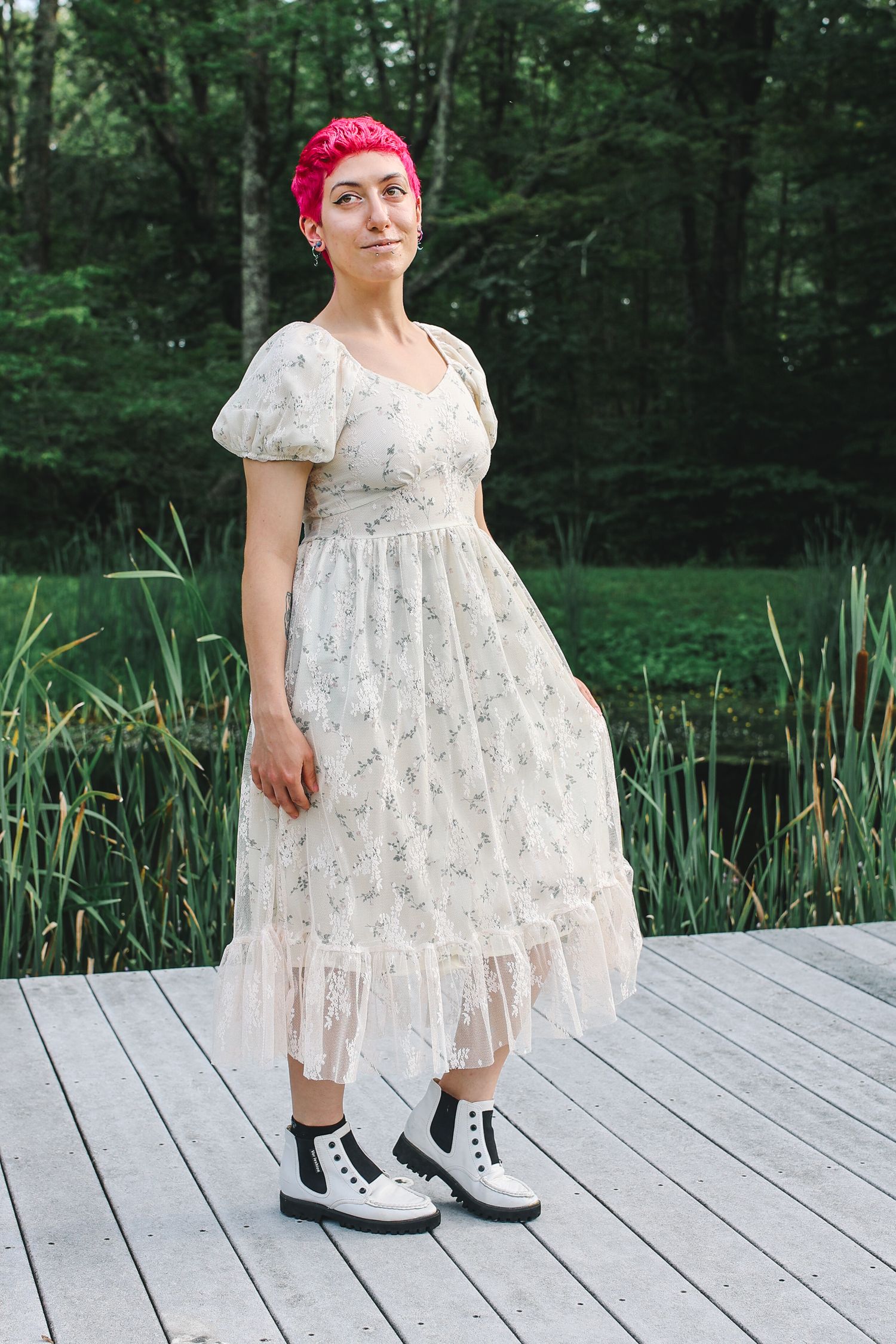 A person in a white lace dress and red headband stands on a wooden deck near a pond surrounded by green forest.