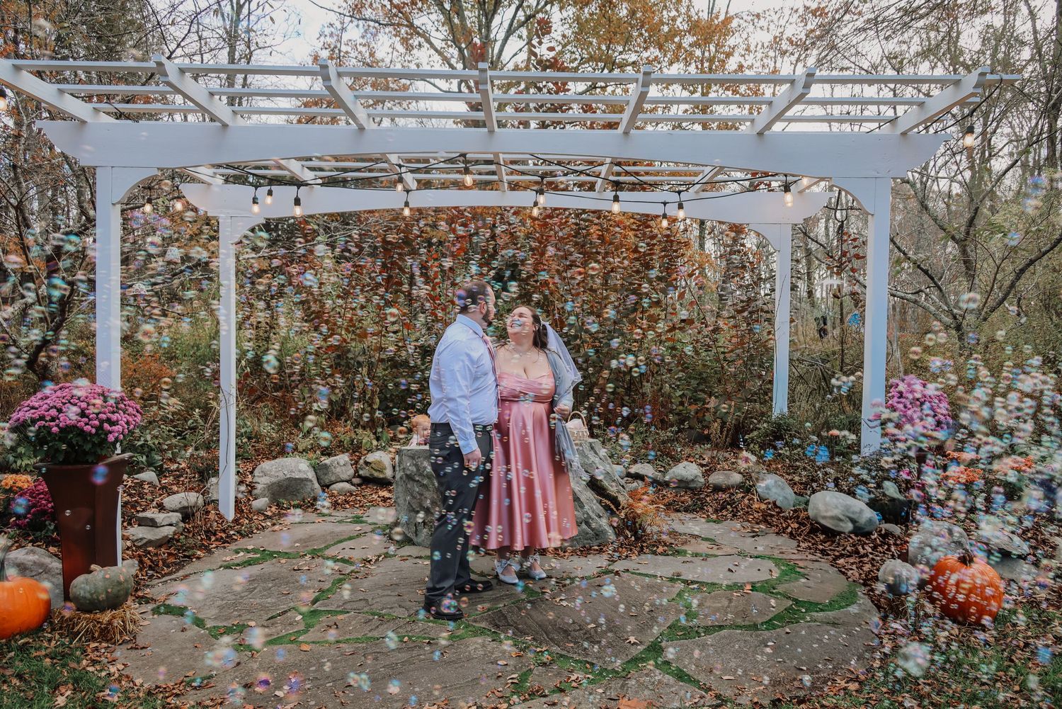 A couple stands beneath a white pergola surrounded by fall decor including pumpkins and colorful flowers.