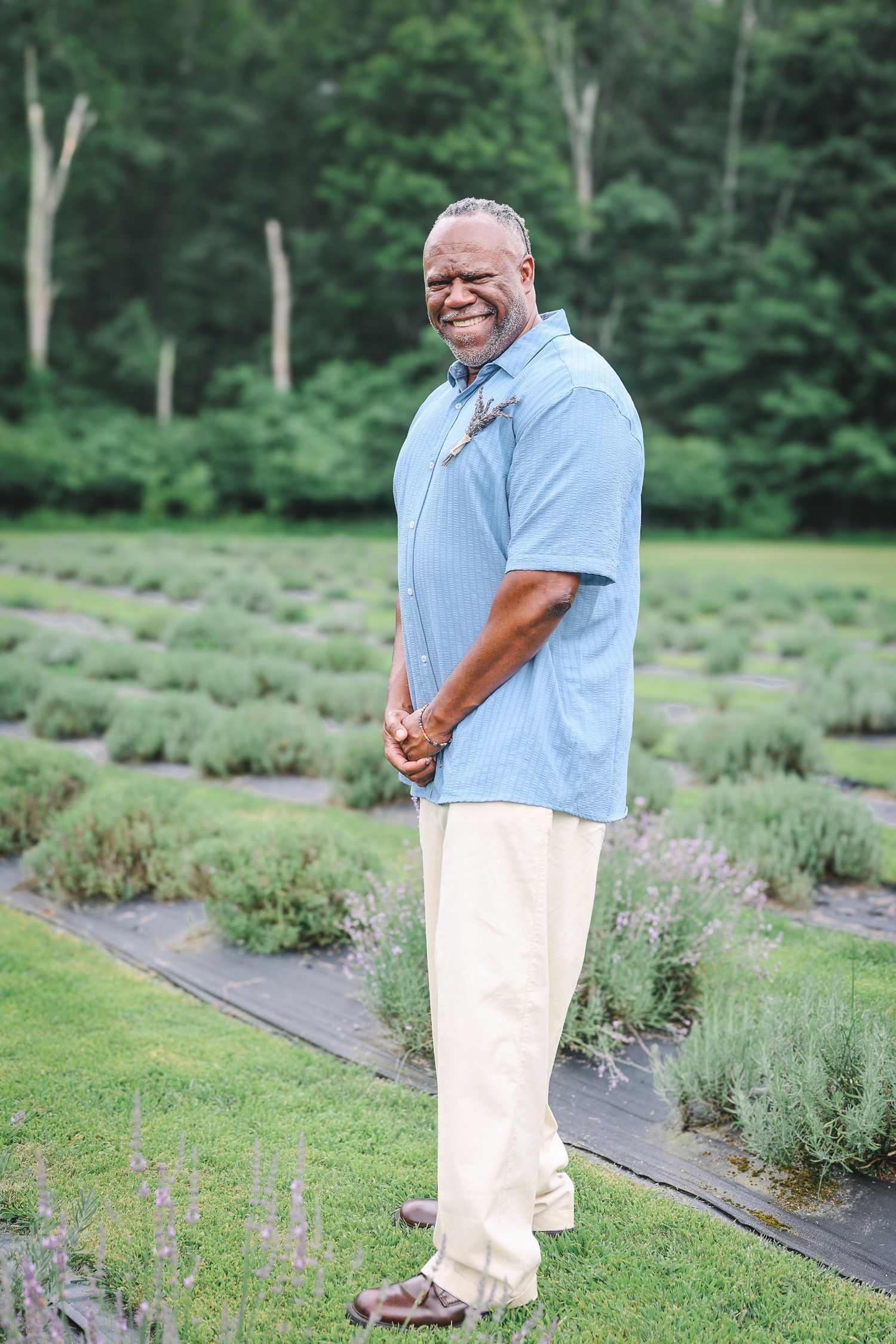 Person in light blue shirt and white pants stands along rows of lavender plants in a garden setting.