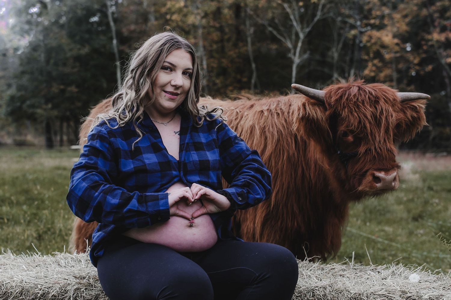 A pregnant woman in a blue plaid flannel shirt makes a heart shape with her hands while standing next to a Highland cow.