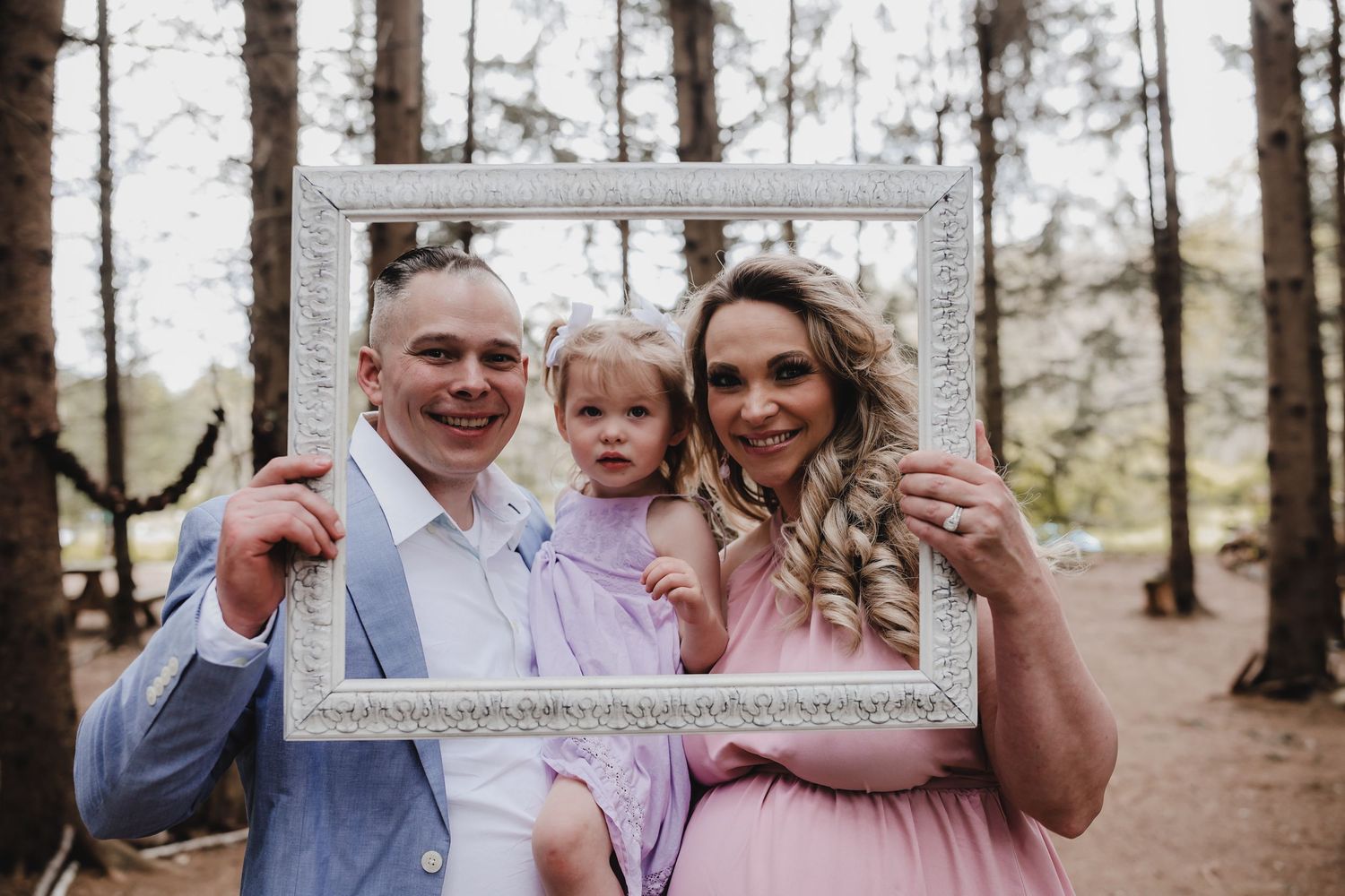 Family poses playfully with white empty picture frame in outdoor forest setting during autumn photoshoot.