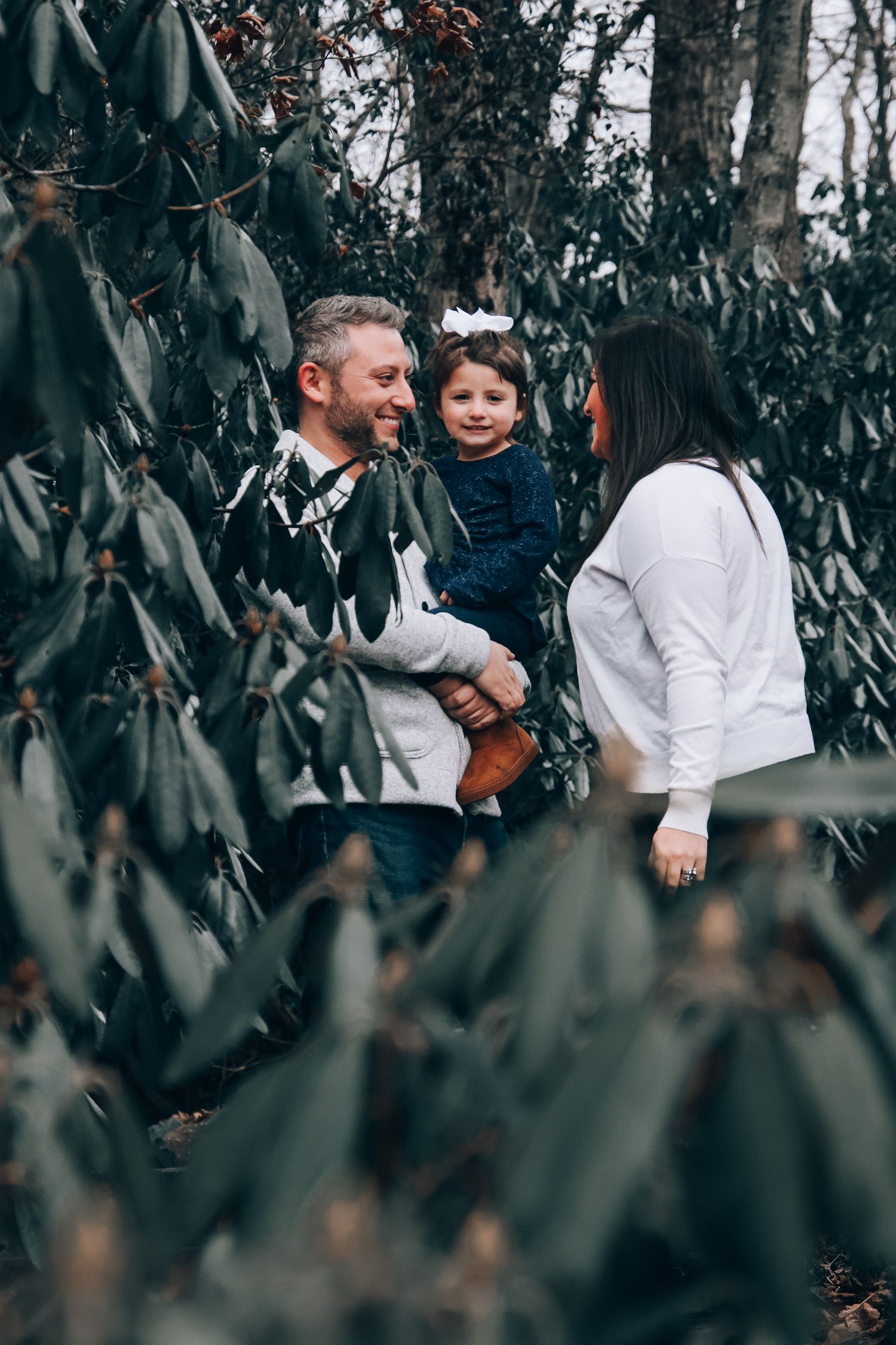Family members share tender moments together while standing among green foliage in a natural outdoor setting.