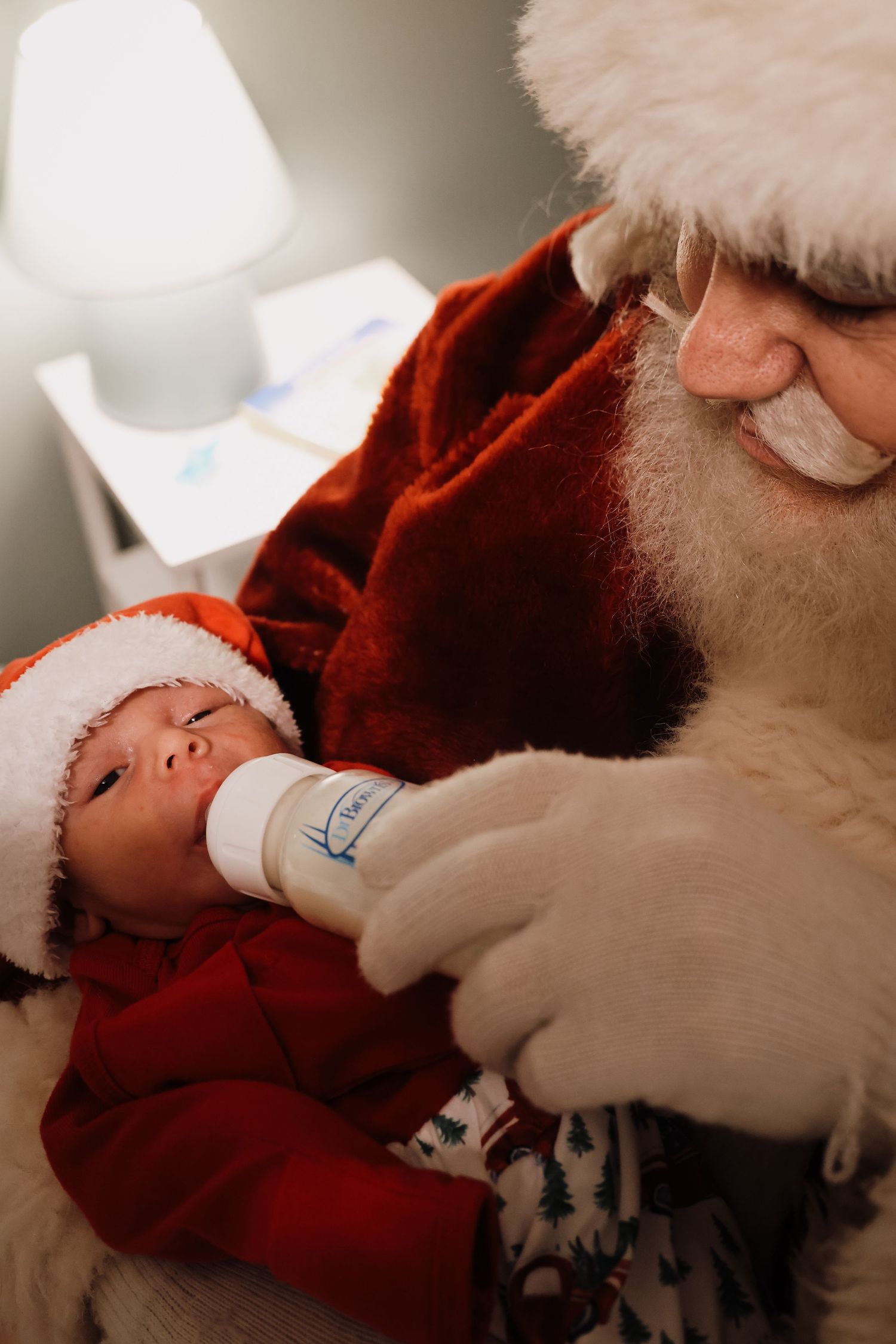 A baby in a red outfit and Santa hat drinks from a bottle while being held in a festive holiday scene.