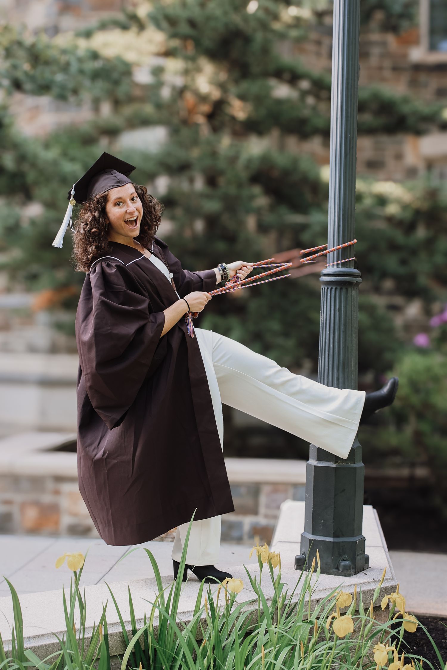 A graduate in cap and gown playfully swings around a lamp post in an outdoor setting with yellow flowers nearby.