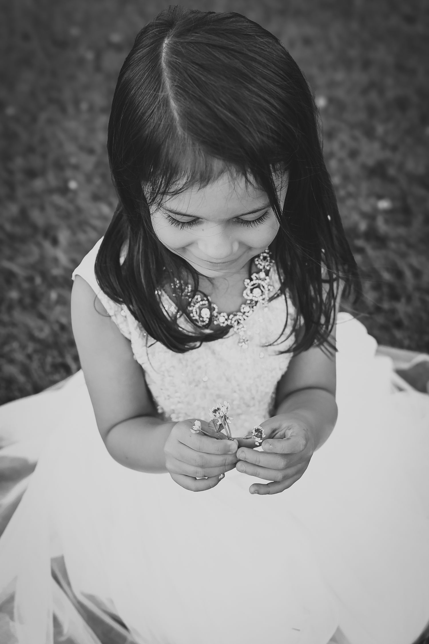 A young girl in a white dress looks down while playing with something in her hands in a black and white photo.