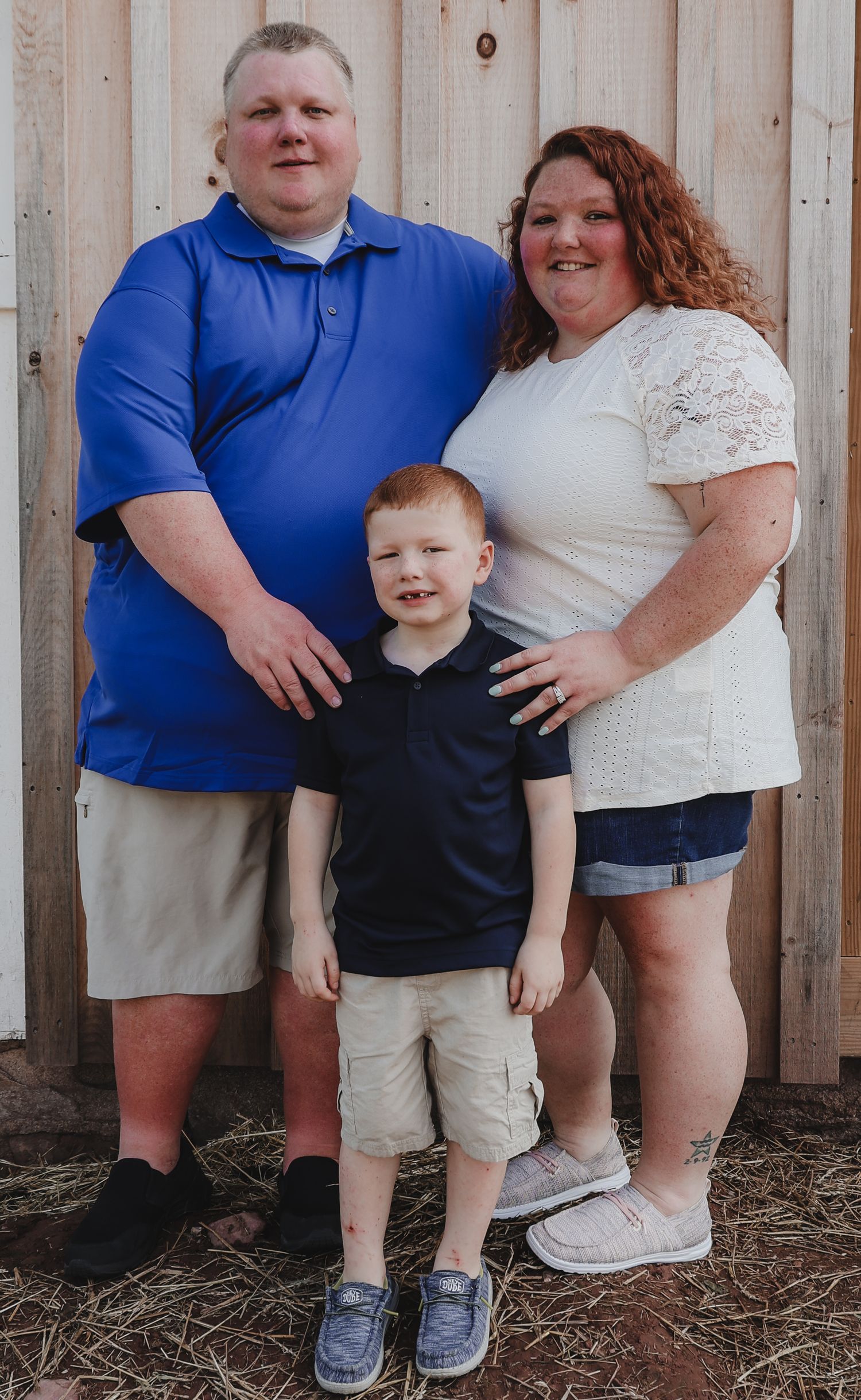 A family of three poses together against a wooden fence background wearing casual summer clothing.