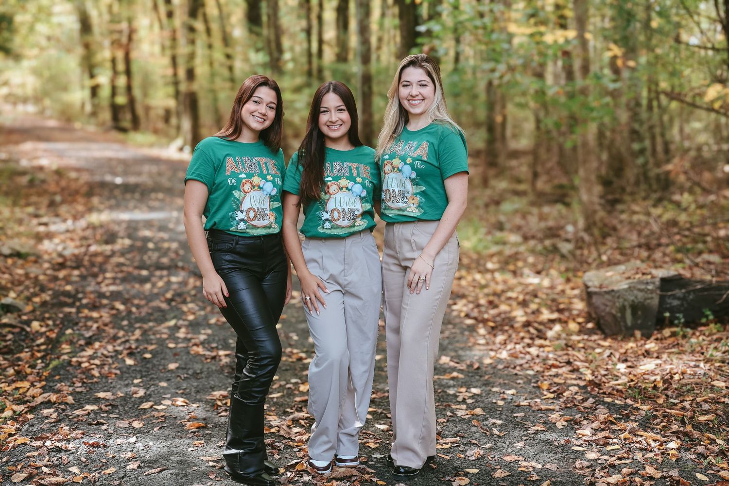 Three people wearing matching green t-shirts stand together smiling in a wooded outdoor setting during autumn.