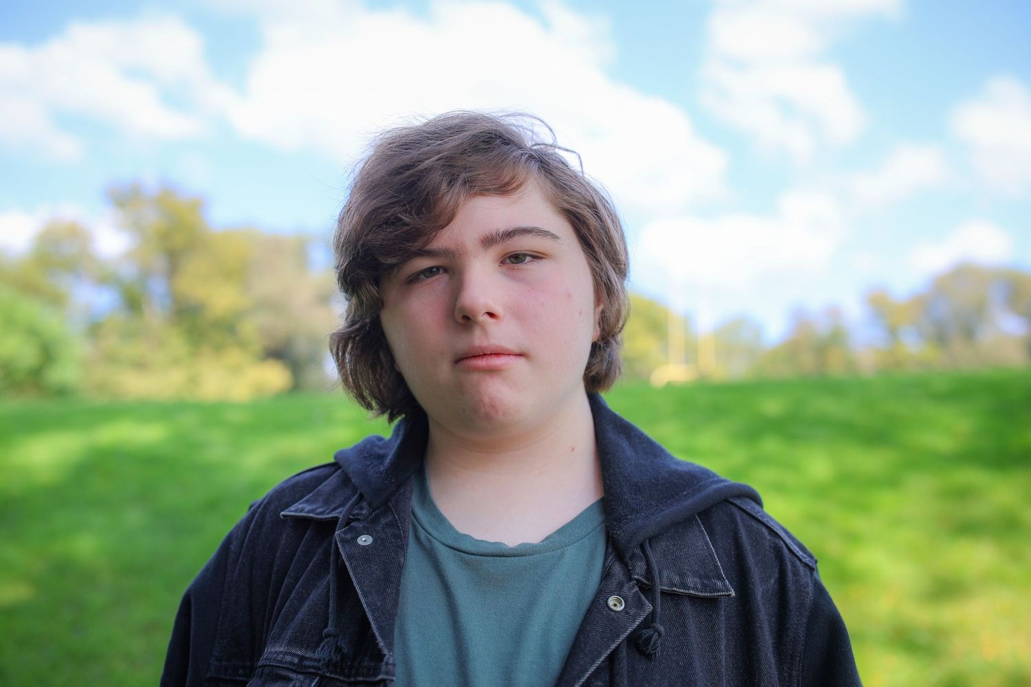 Person wearing a dark jacket stands in front of a green grassy hillside on a sunny day.