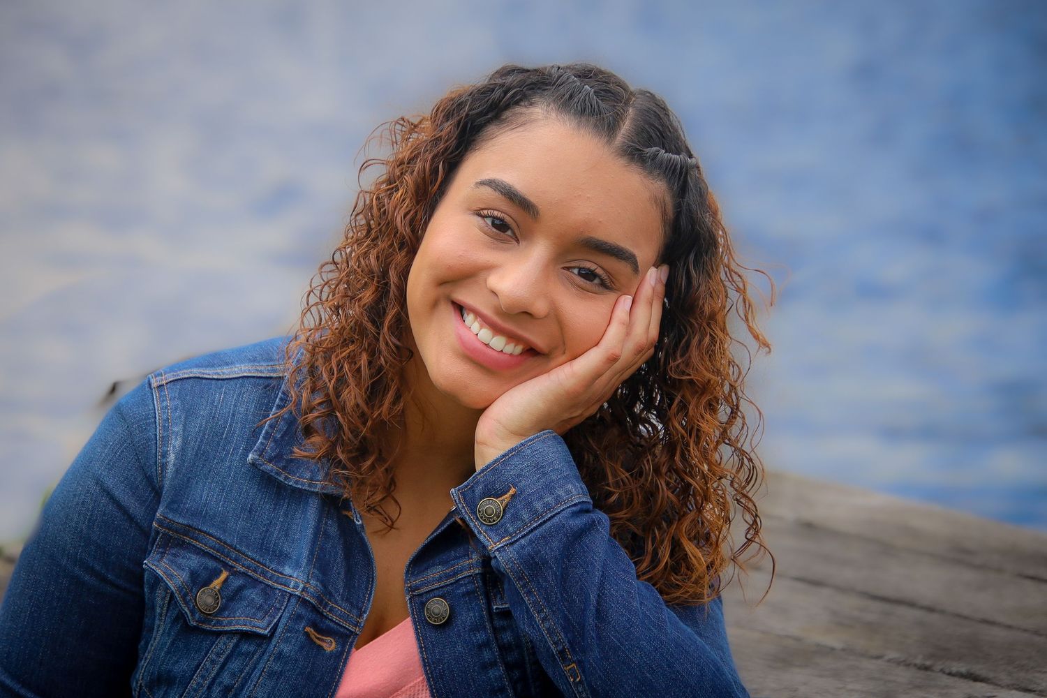 A person with curly hair and denim jacket smiles warmly while posing against a cloudy blue sky backdrop.