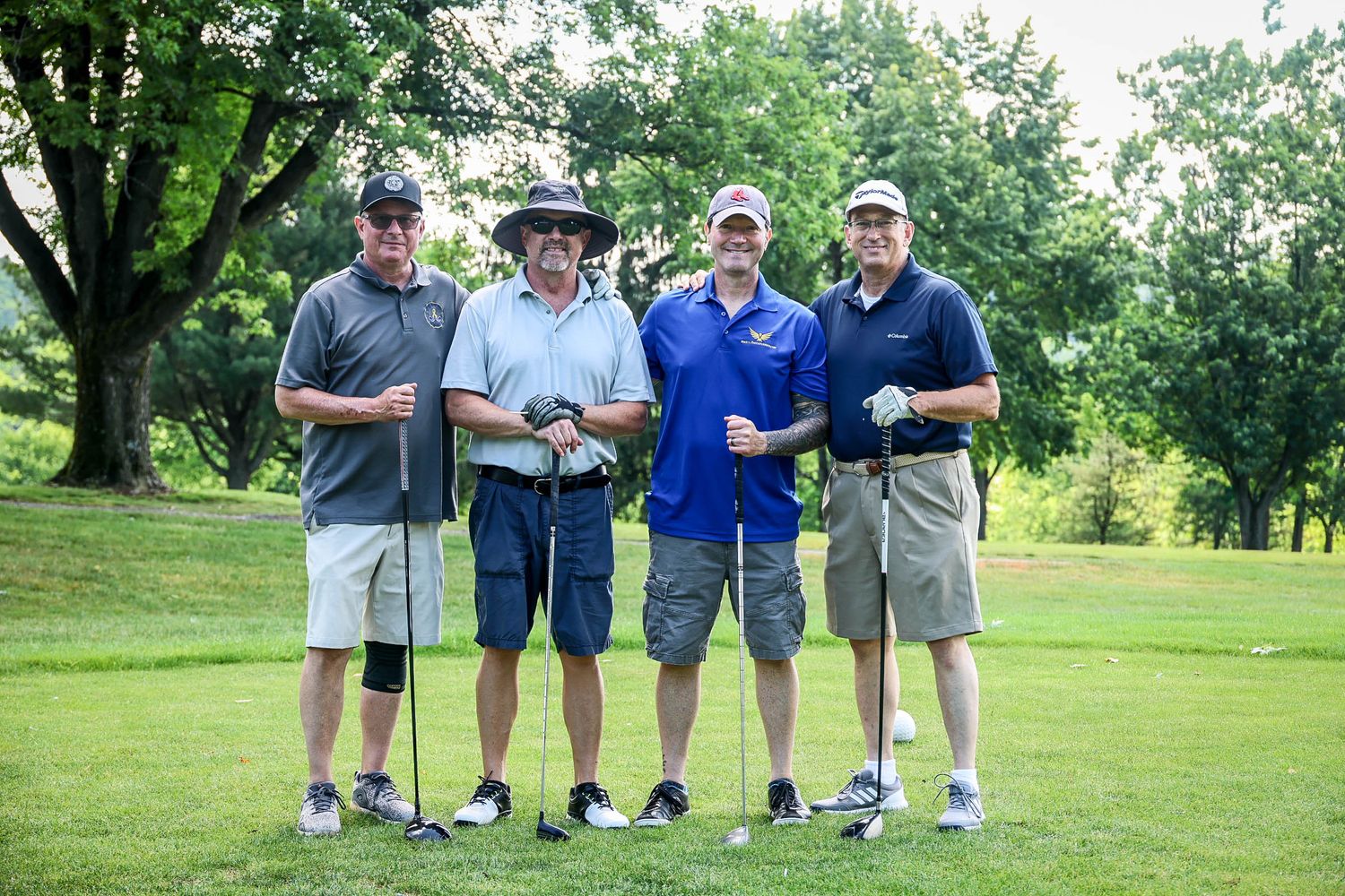 Four men posing together on a golf course, holding clubs, surrounded by lush green trees and fairway.
