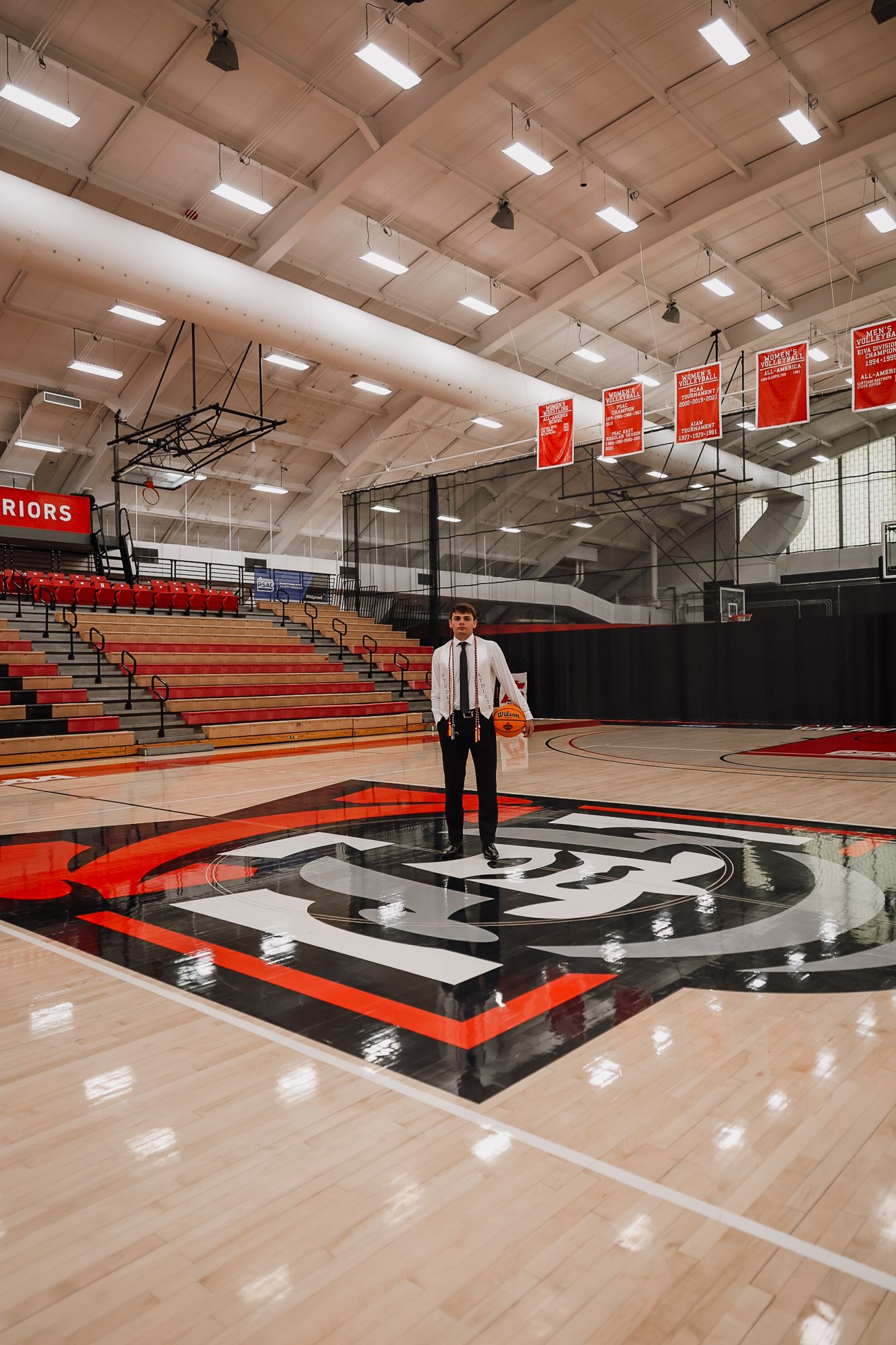 Person standing at center court in an empty Warriors basketball gymnasium holding a ball, wearing formal attire.