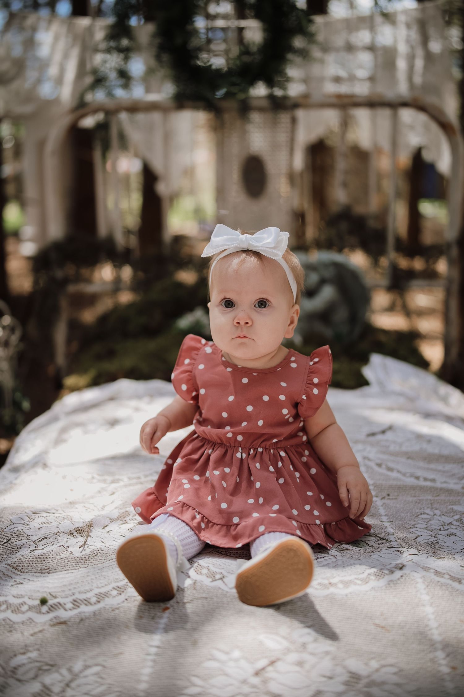 Baby girl in pink polka dot dress and white bow headband sitting on lace blanket outdoors.
