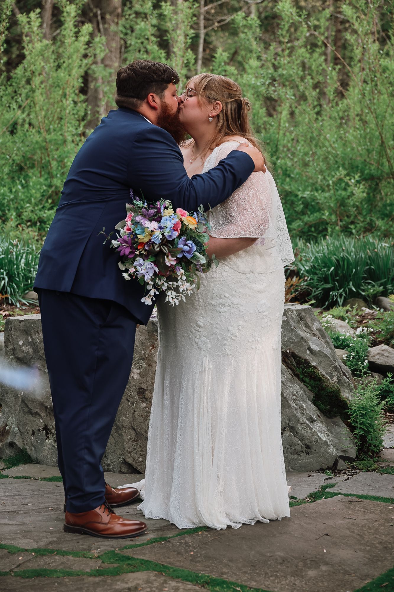 Bride and groom share first kiss in outdoor garden wedding ceremony, bride holds colorful wildflower bouquet, groom in navy suit.