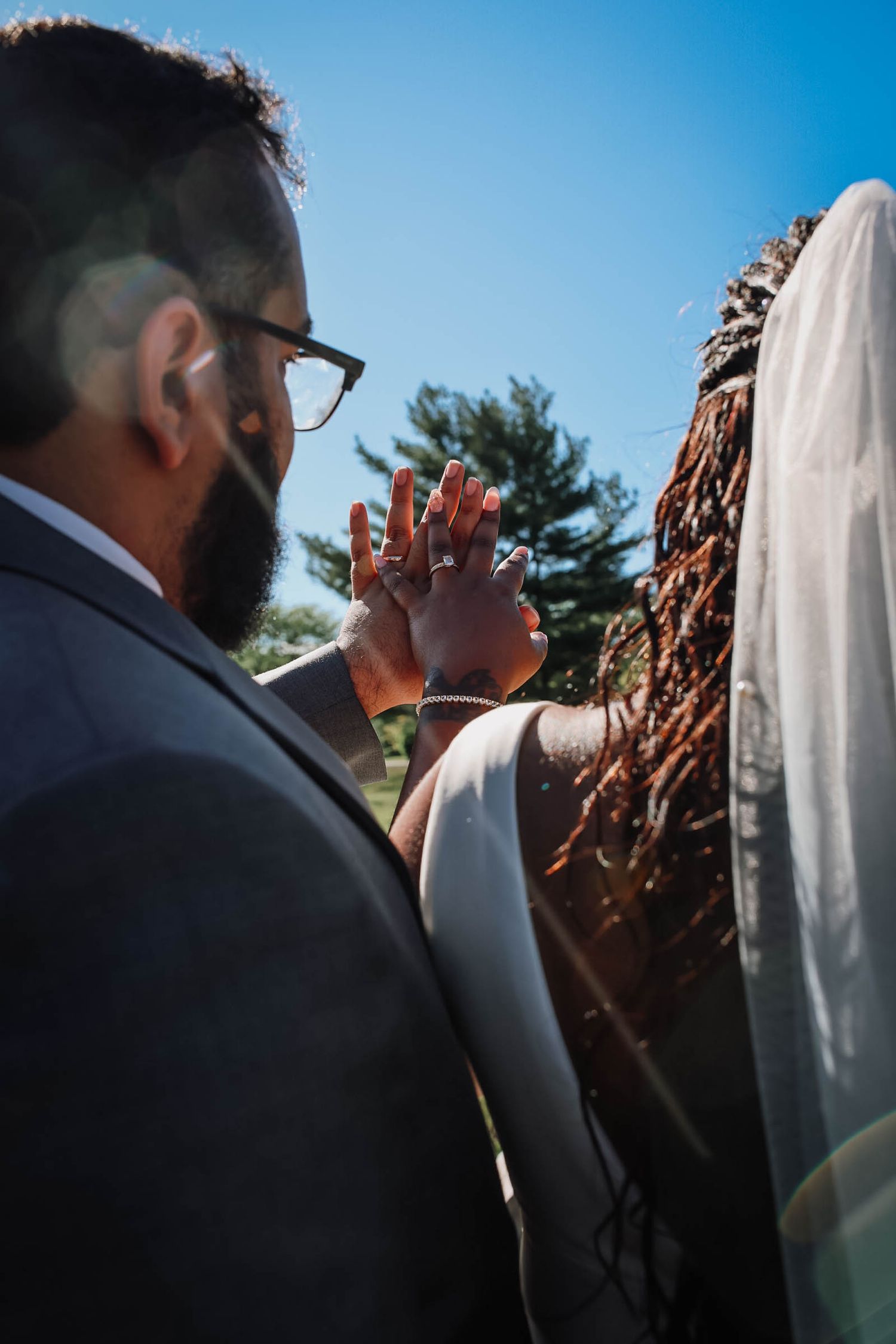 Newlywed couple holding hands showing wedding rings outdoors on a sunny day, groom in gray suit with bride in veil.