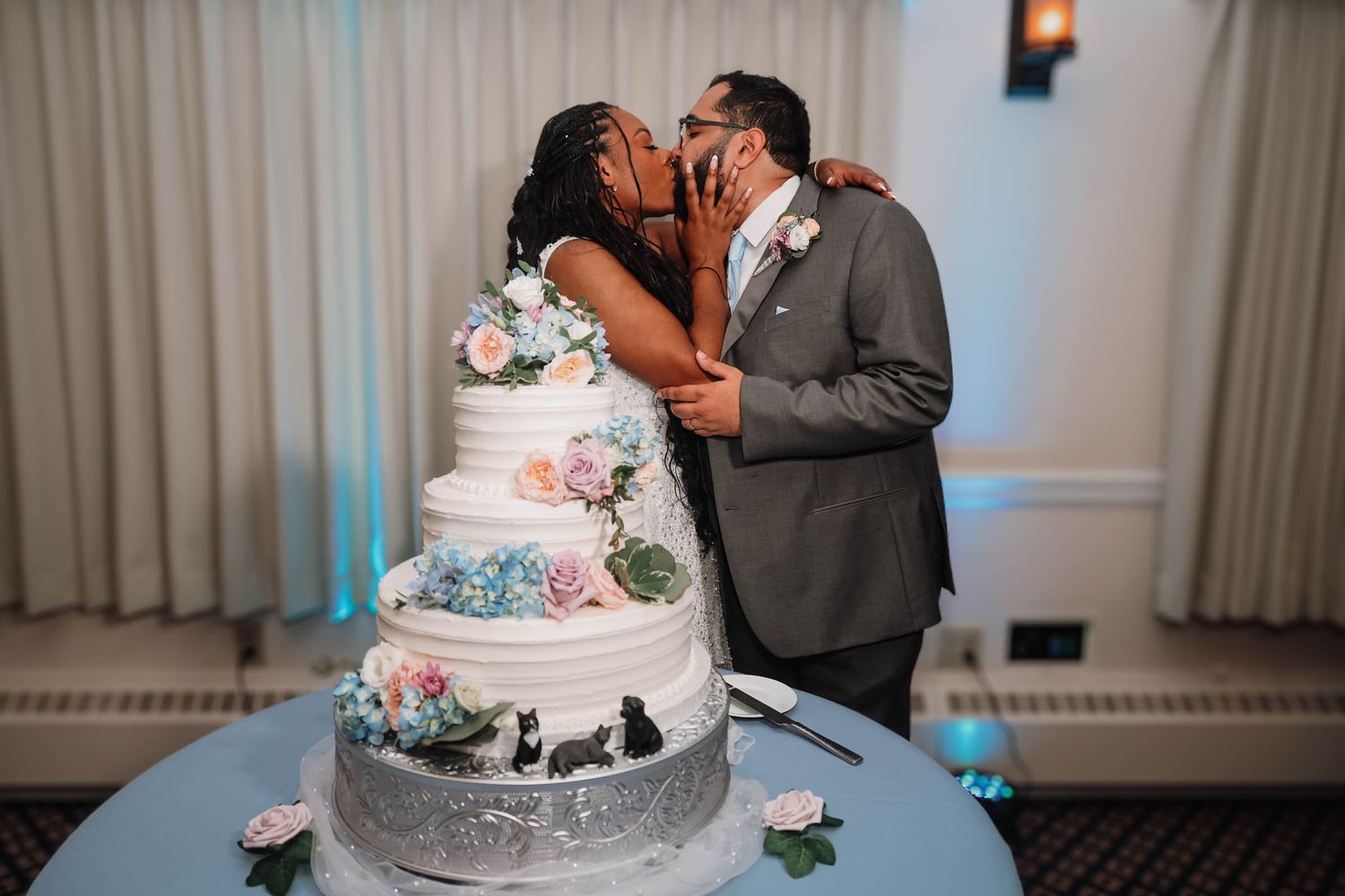 Newlywed couple sharing a kiss behind a elegant four-tier white wedding cake decorated with colorful flowers.