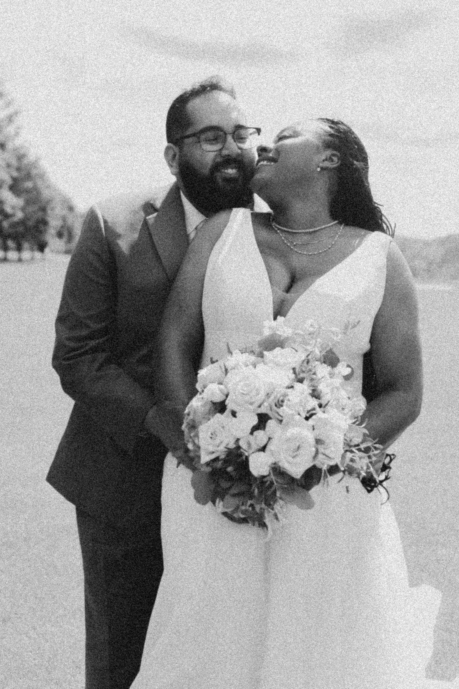 Black and white photo of a smiling bride and groom in wedding attire, bride holding a white floral bouquet.