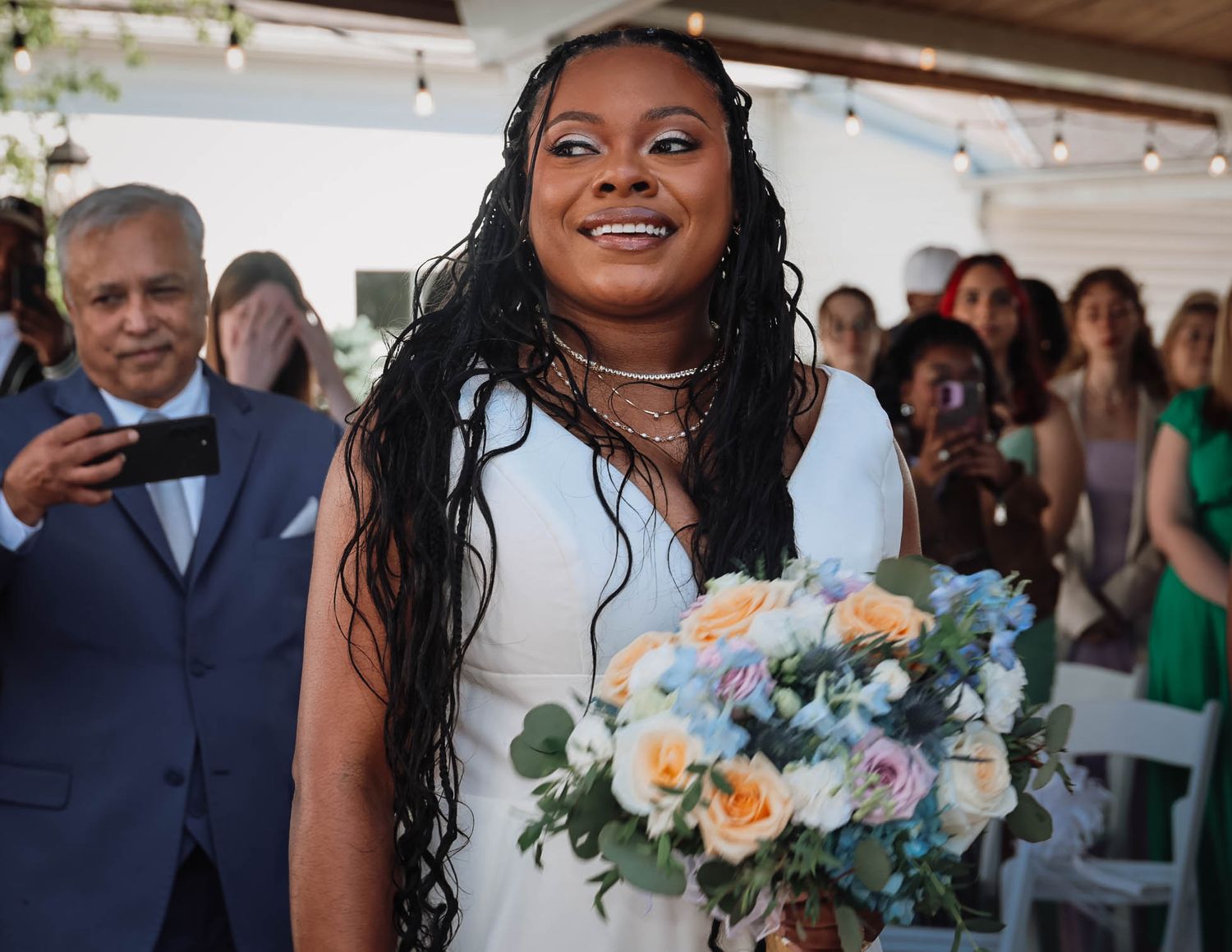 A smiling bride in a white dress holds a pastel bouquet while walking down the aisle at an outdoor wedding ceremony.