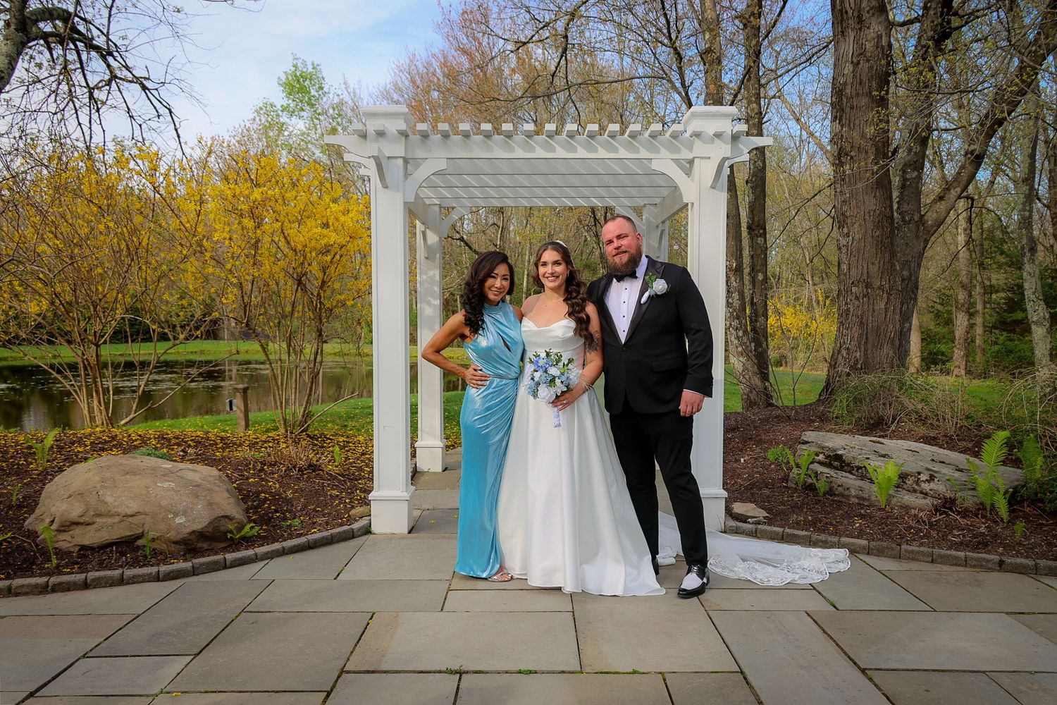 Bride and groom pose with a guest under a white pergola in a lush garden setting during an outdoor wedding ceremony.