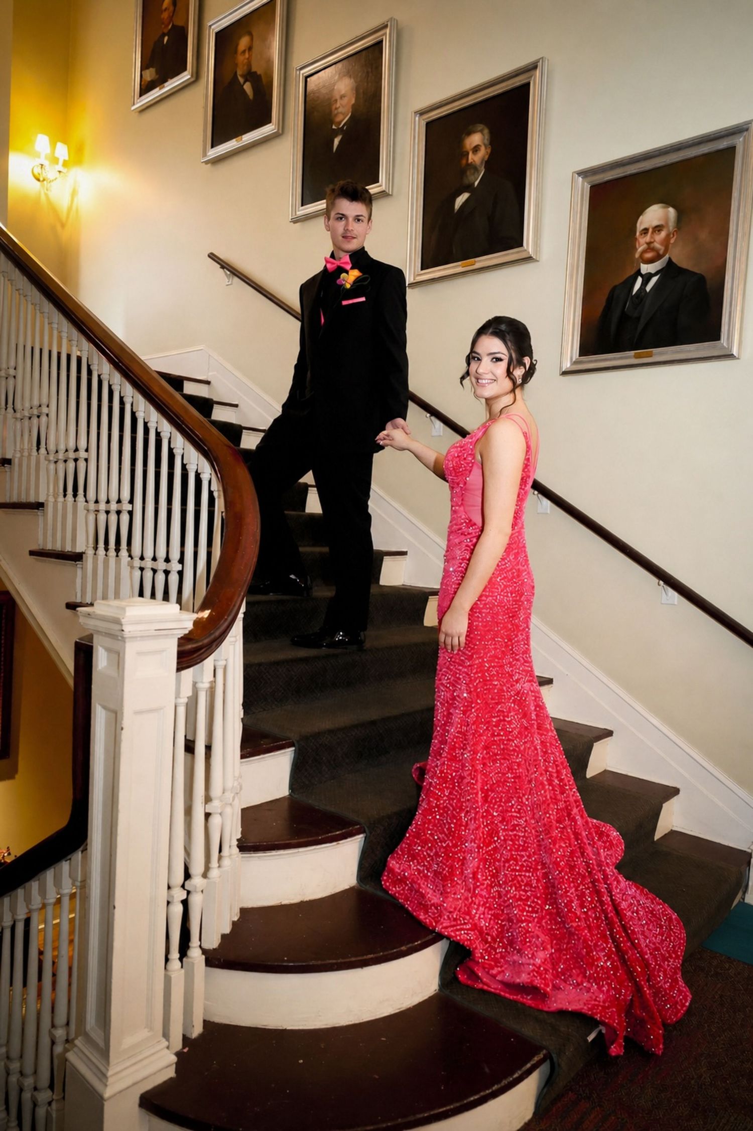 Couple in formal wear on elegant staircase, woman in red gown, man in black tuxedo, portrait paintings on wall.