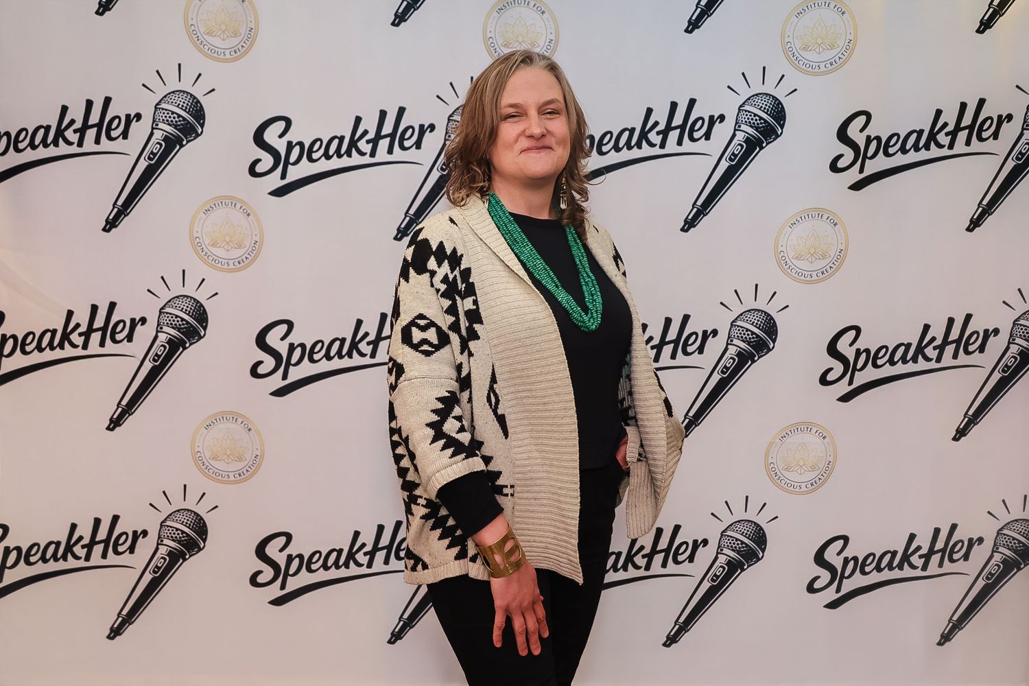 Woman in patterned cardigan posing at SpeakHer event backdrop with microphone logos.