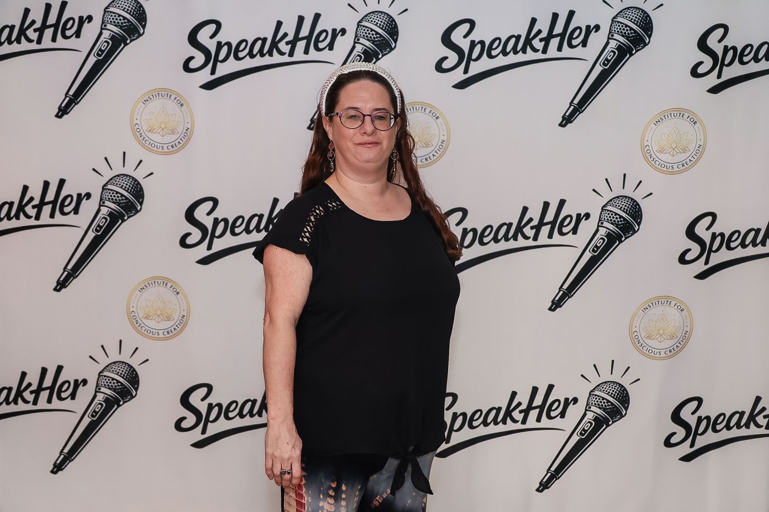 Woman in black top posing at SpeakHer event backdrop with microphone logo branding.