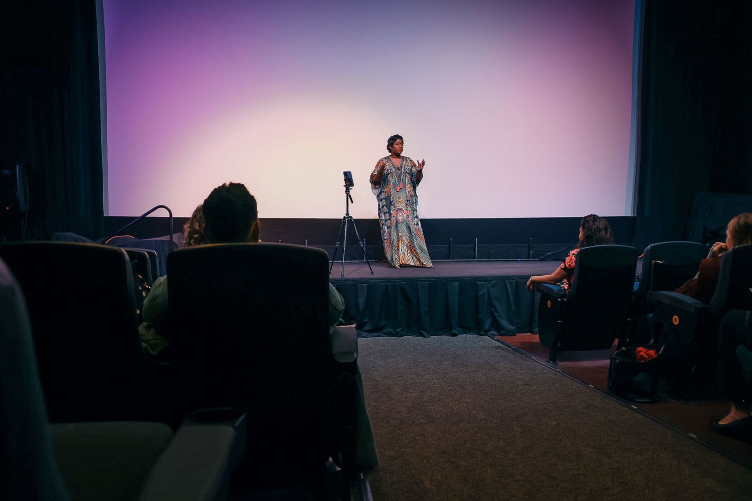Performer in elegant gown speaking on stage before small audience in dimly lit theater with large screen backdrop.