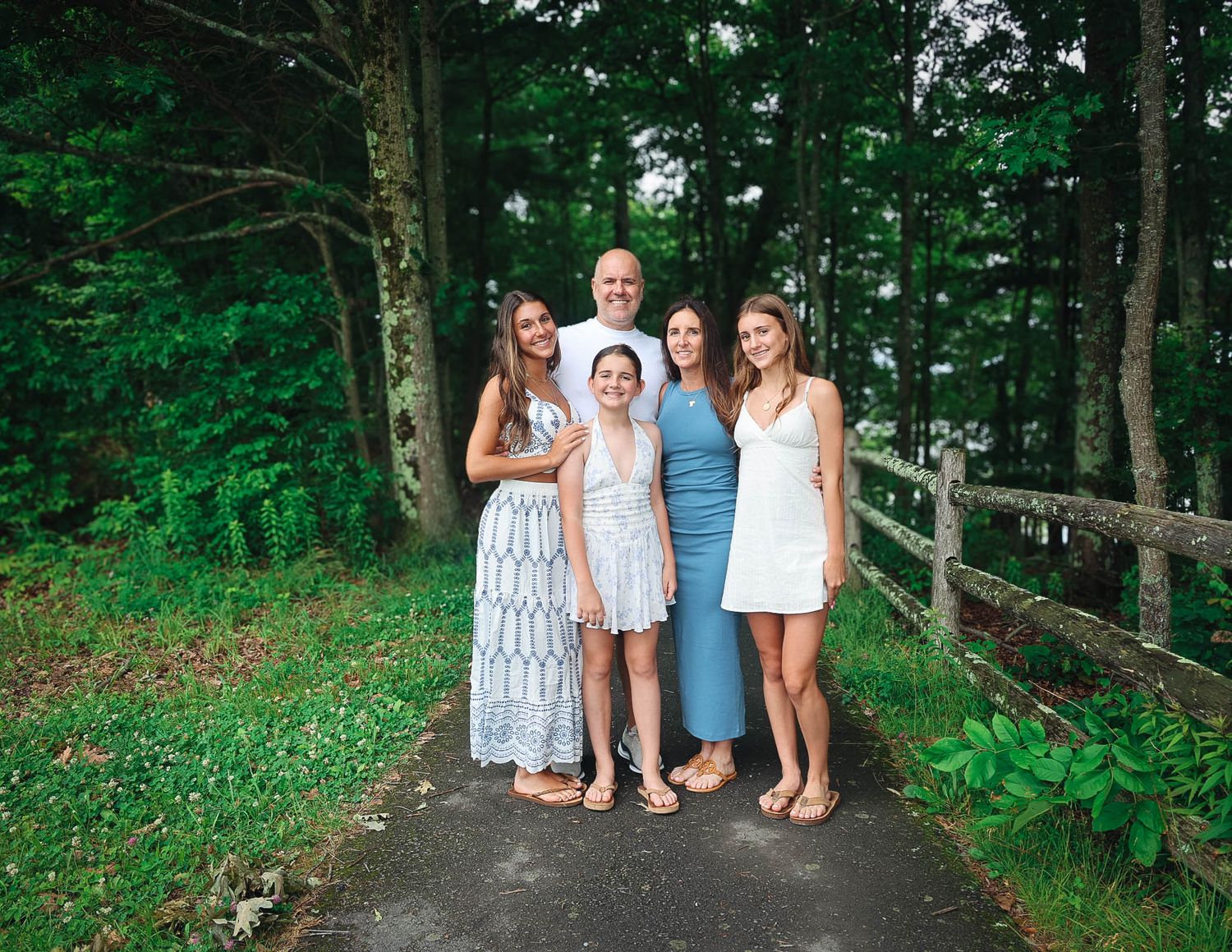 Family of five smiling together on a wooded path with lush green trees and a wooden fence in the background.