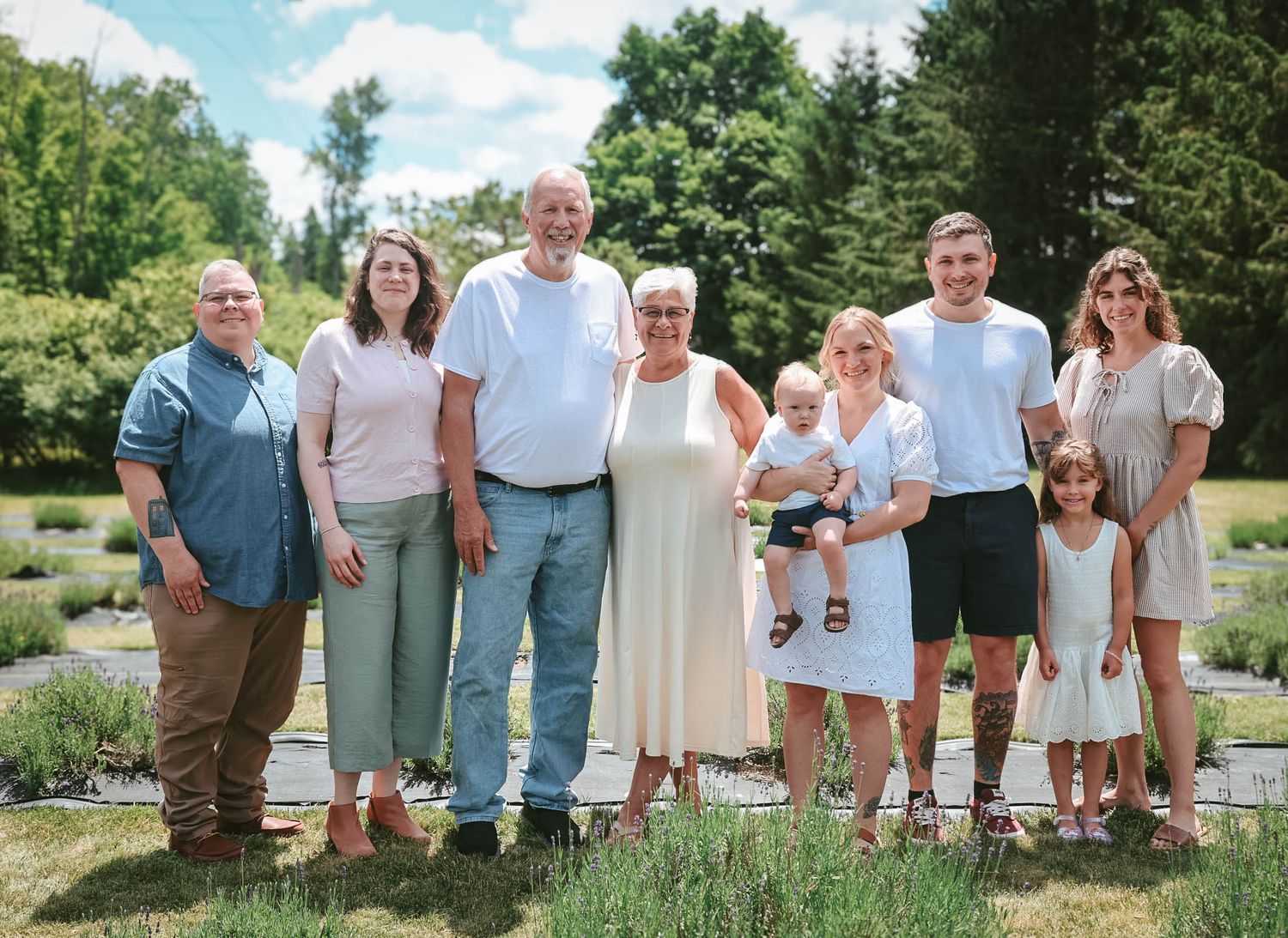 A multigenerational family of eight poses together outdoors in a sunny garden setting with lush green trees in the background.