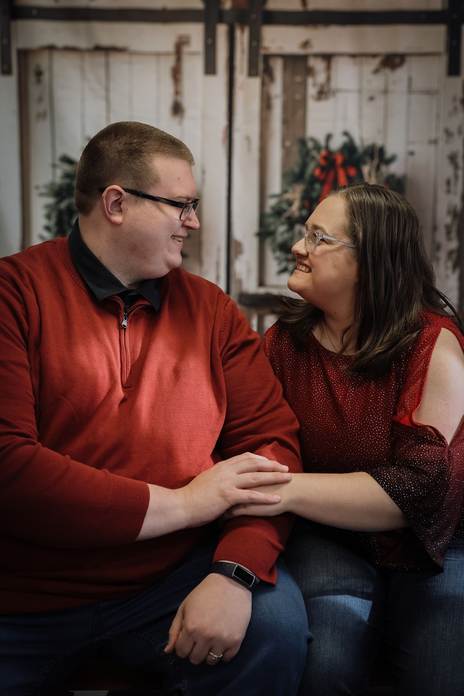 Couple in red sweaters holding hands and smiling at each other in front of a rustic white barn door with holiday wreath.