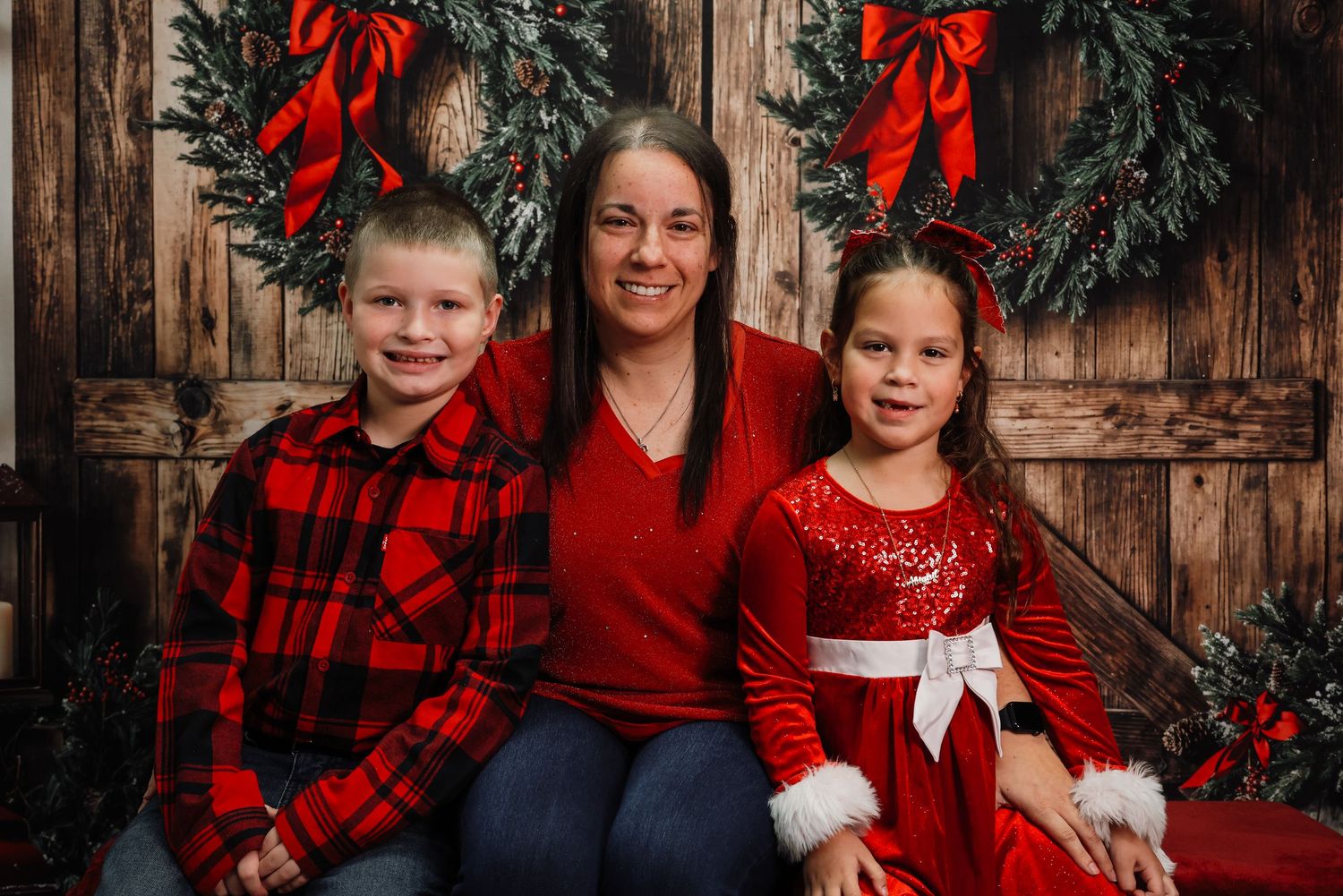 Family of three posing for Christmas photo in red outfits against rustic wood backdrop with holiday wreaths.