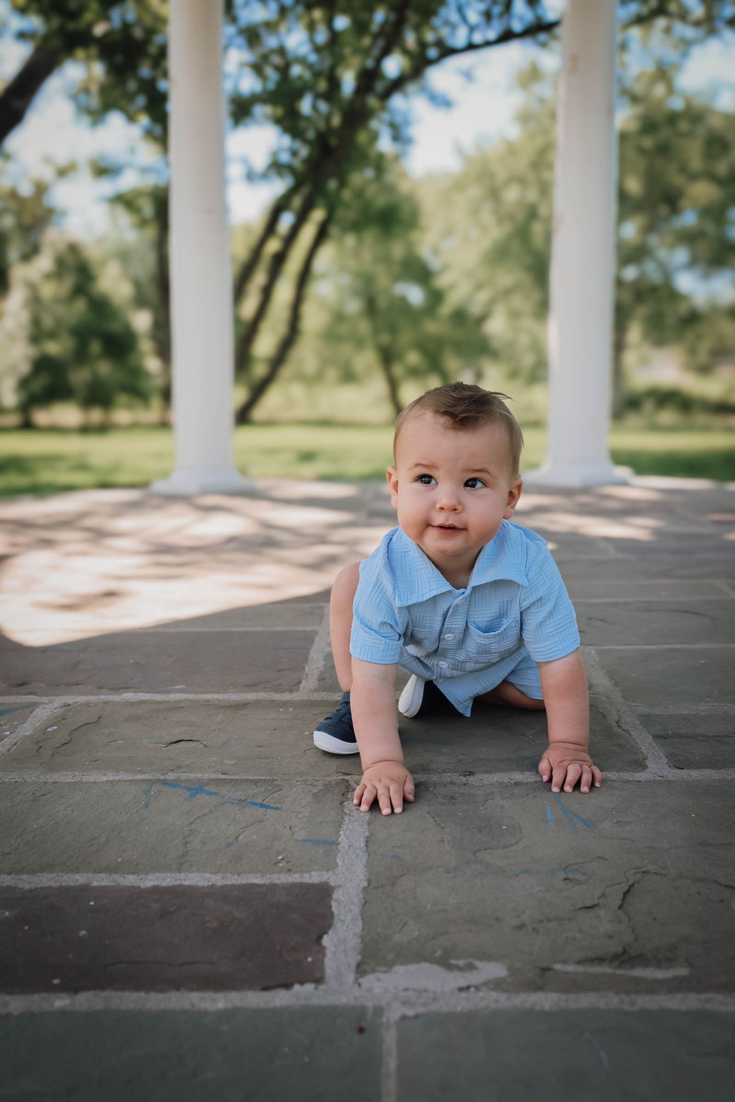 A baby boy in a blue polo romper crawls on a stone patio surrounded by white columns and green trees.