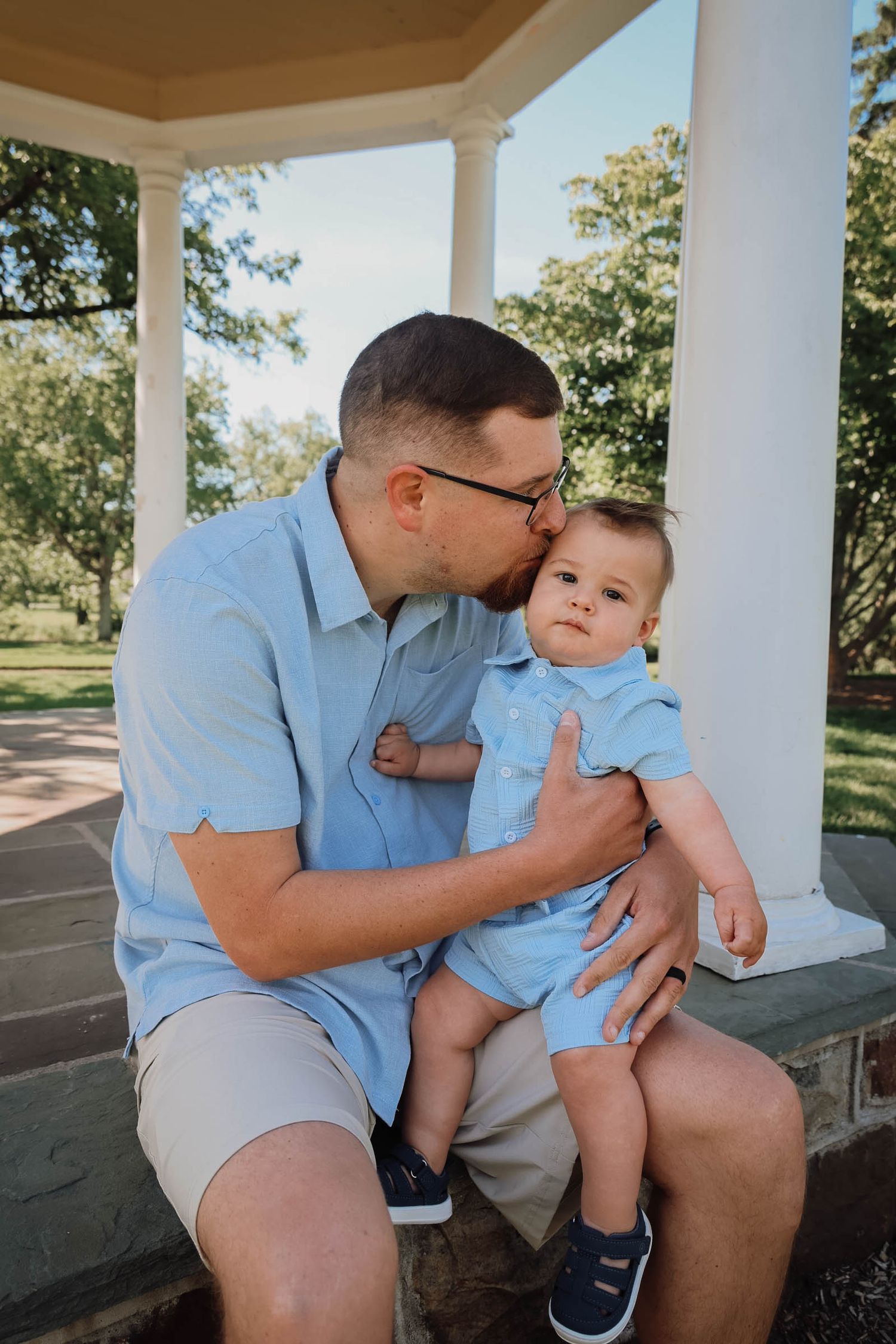 A father in a blue shirt kisses his baby boy on a gazebo steps in a sunny park.