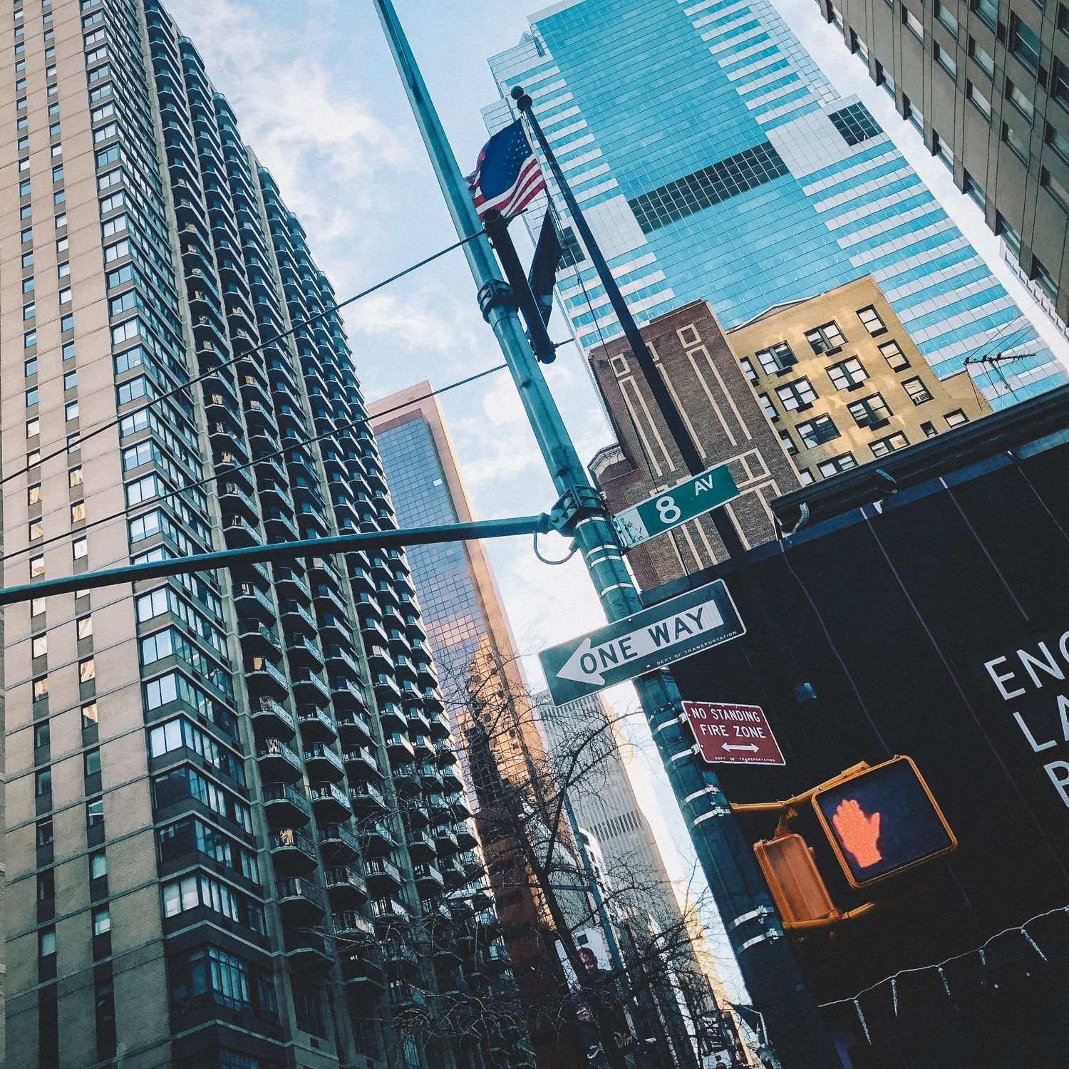 Low-angle view of NYC street intersection with One Way sign, traffic light, American flag, and modern skyscrapers against blue sky.