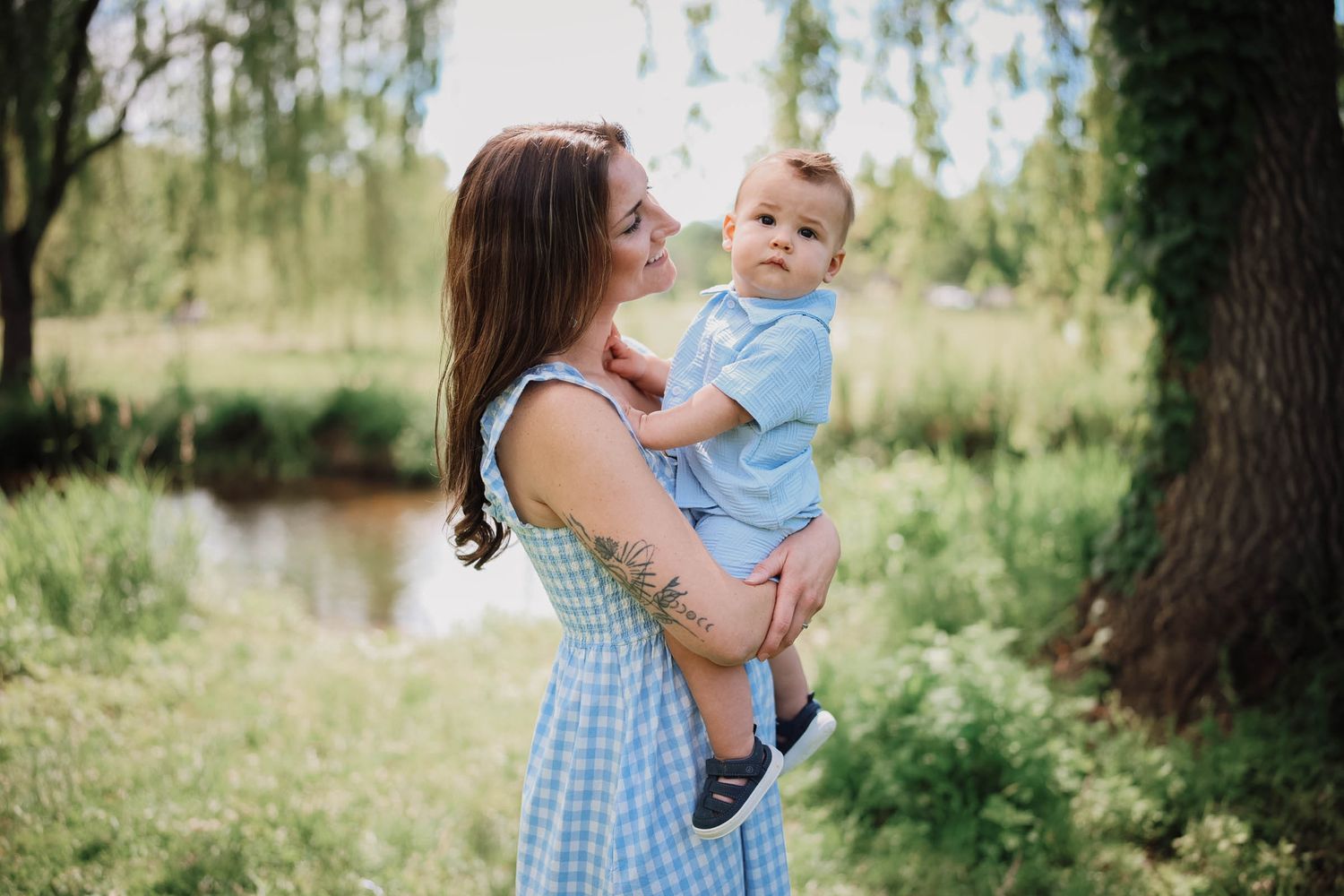 A mother in a blue gingham dress holds her baby boy outdoors near a pond surrounded by lush green trees.