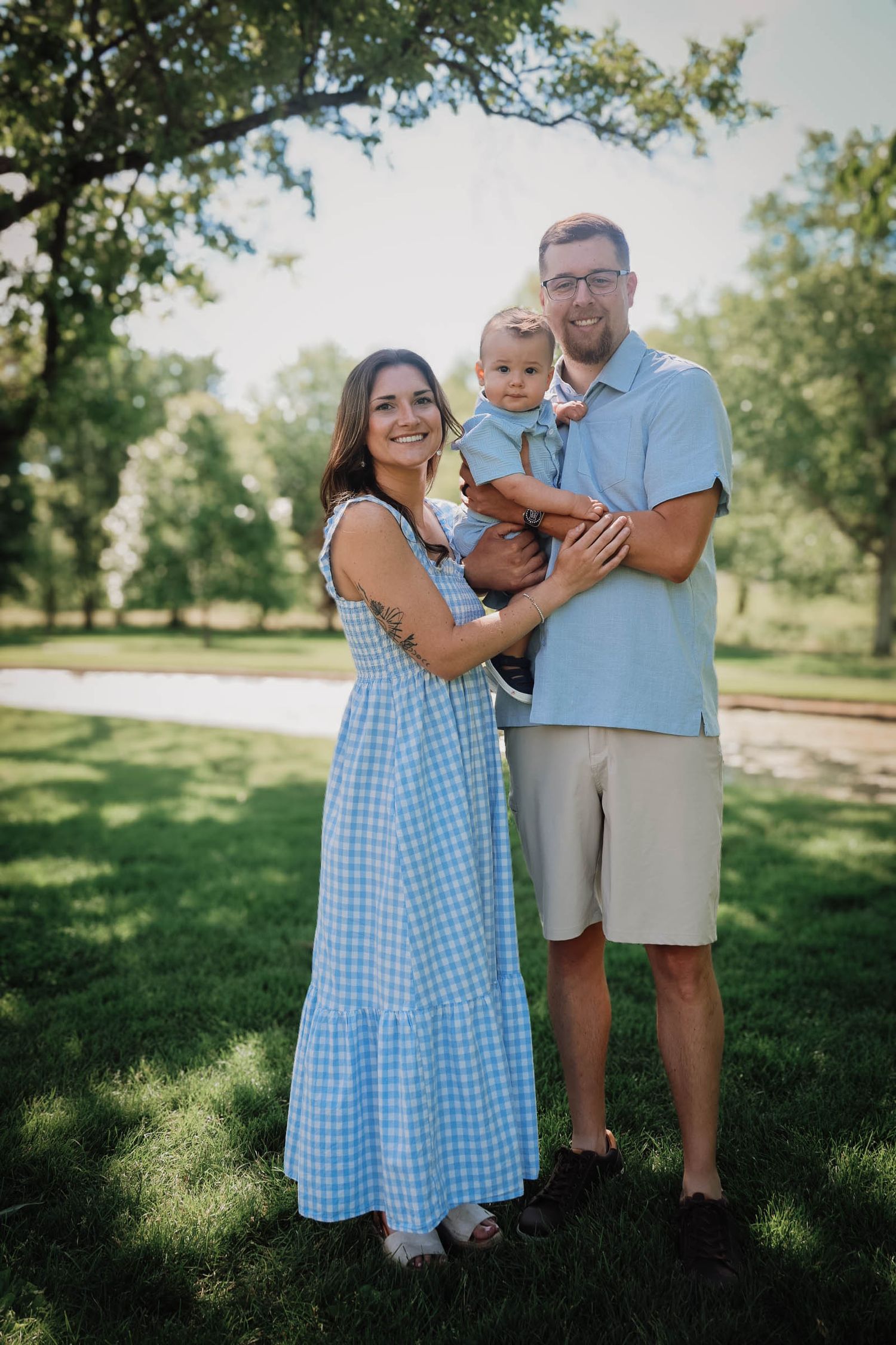 Happy young family with baby posing outdoors in green park, mother in blue gingham dress, father in light blue shirt.