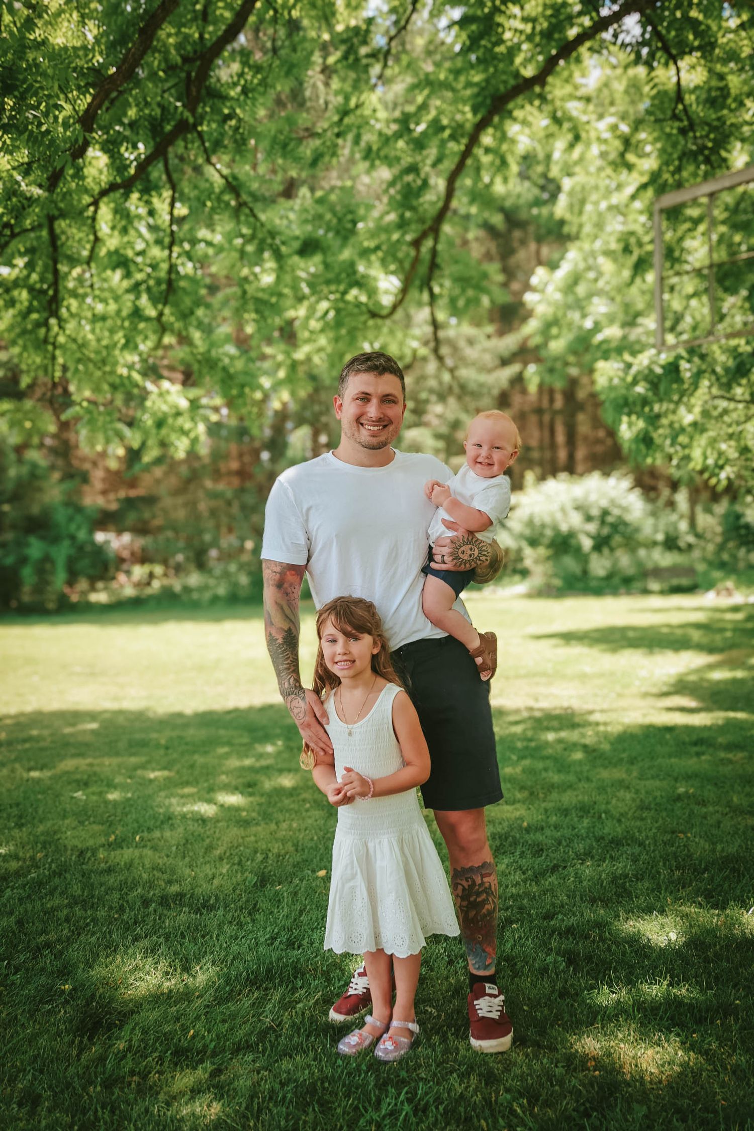 Father with tattoos holding baby while young girl in white dress stands beside him on lush green lawn outdoors.