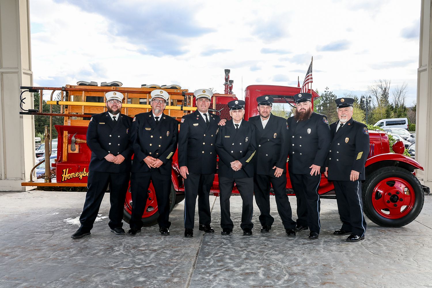 Seven firefighters in dress uniforms pose proudly in front of a vintage red fire truck outside a fire station.