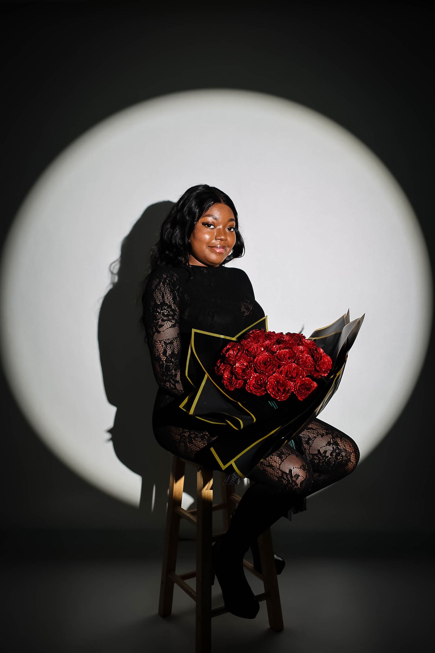 Woman in black lace outfit sitting on stool holding red roses bouquet, illuminated by spotlight on dark background.