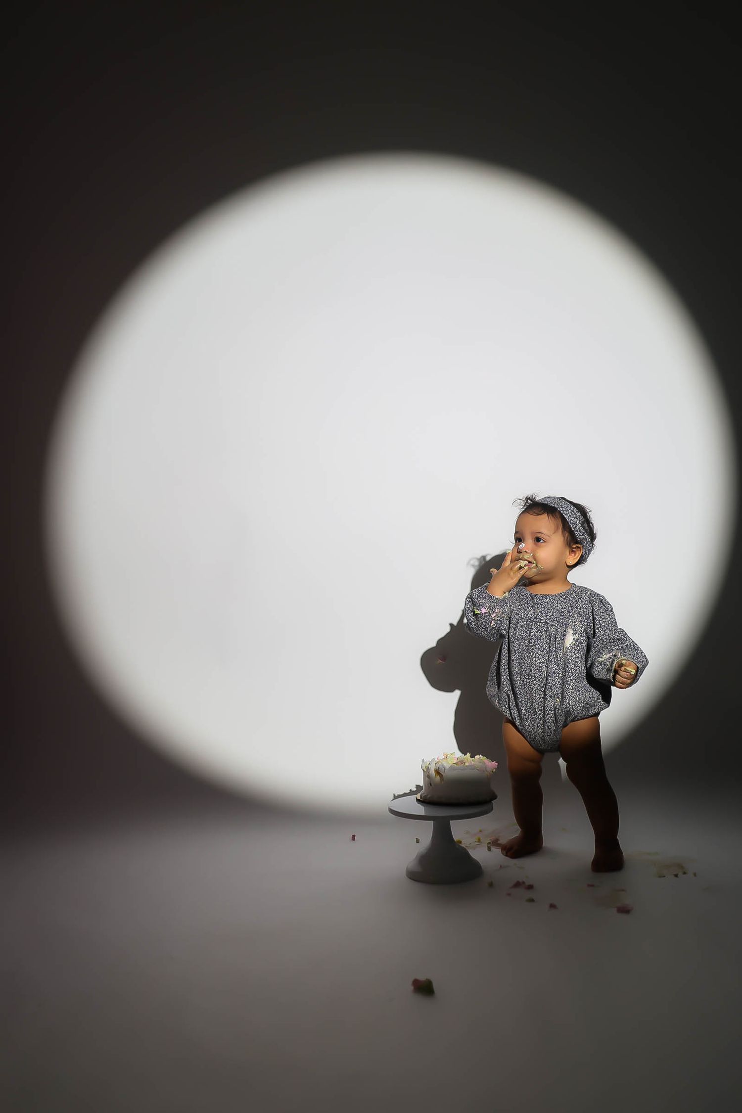 Toddler in gray romper eating cake with a small white cake on a stand under a circular studio spotlight.