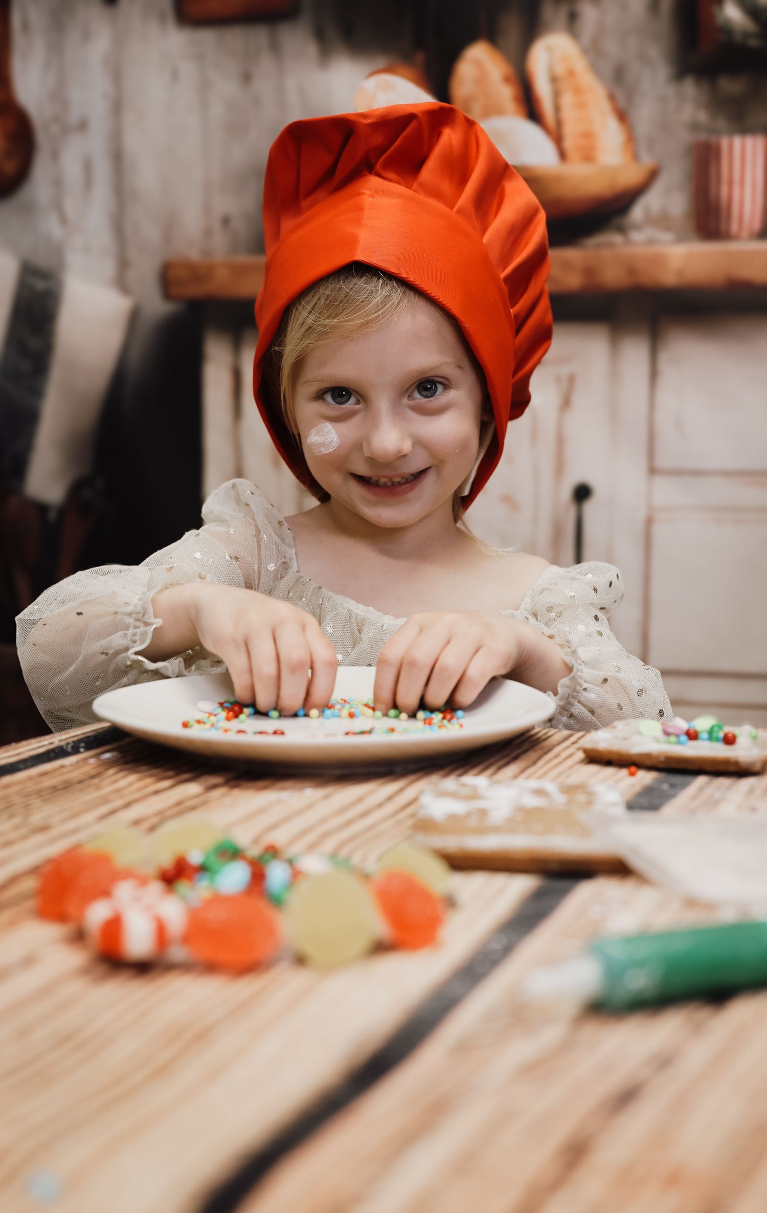 A smiling child wearing an orange chef hat decorates cookies with colorful sprinkles and candies on a wooden table.