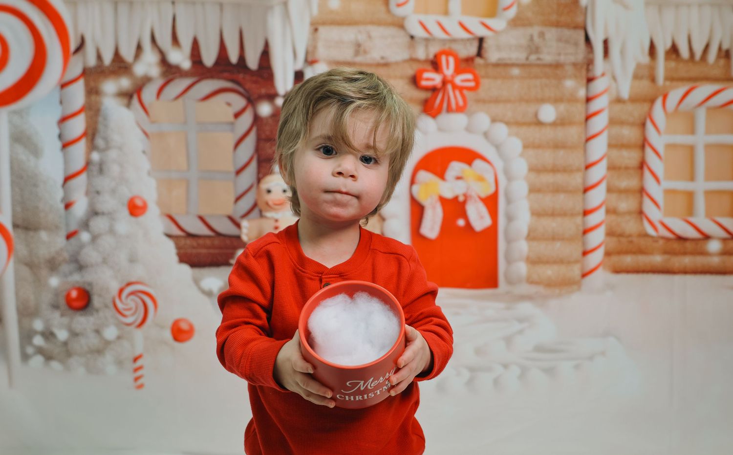 A toddler in a red outfit holds a red bucket with white fluff in front of a festive gingerbread house backdrop.