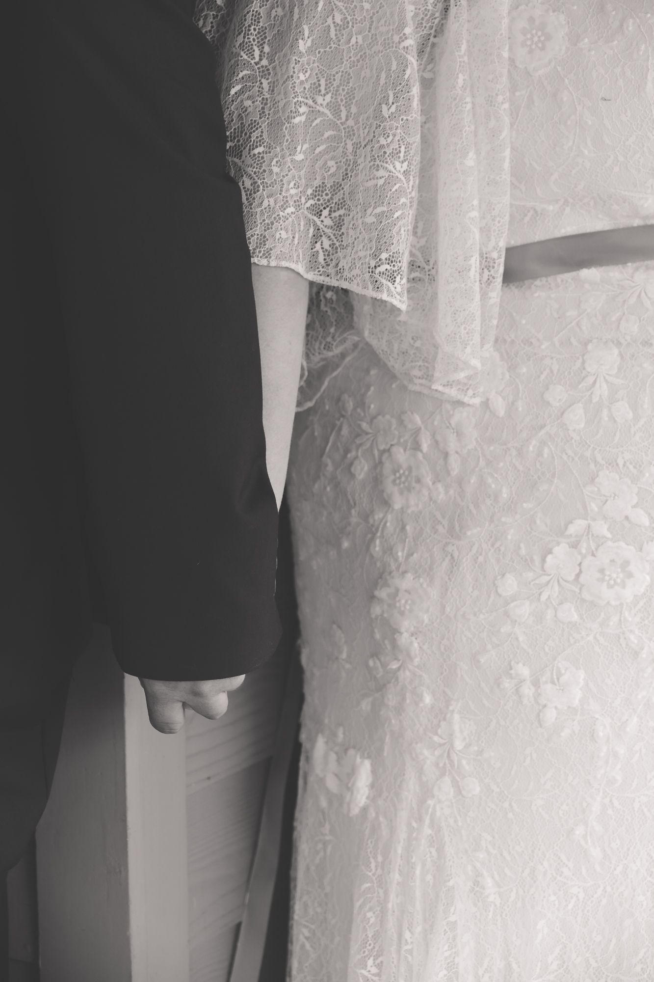 Black and white photo of bride and groom holding hands, showcasing lace wedding dress detail.