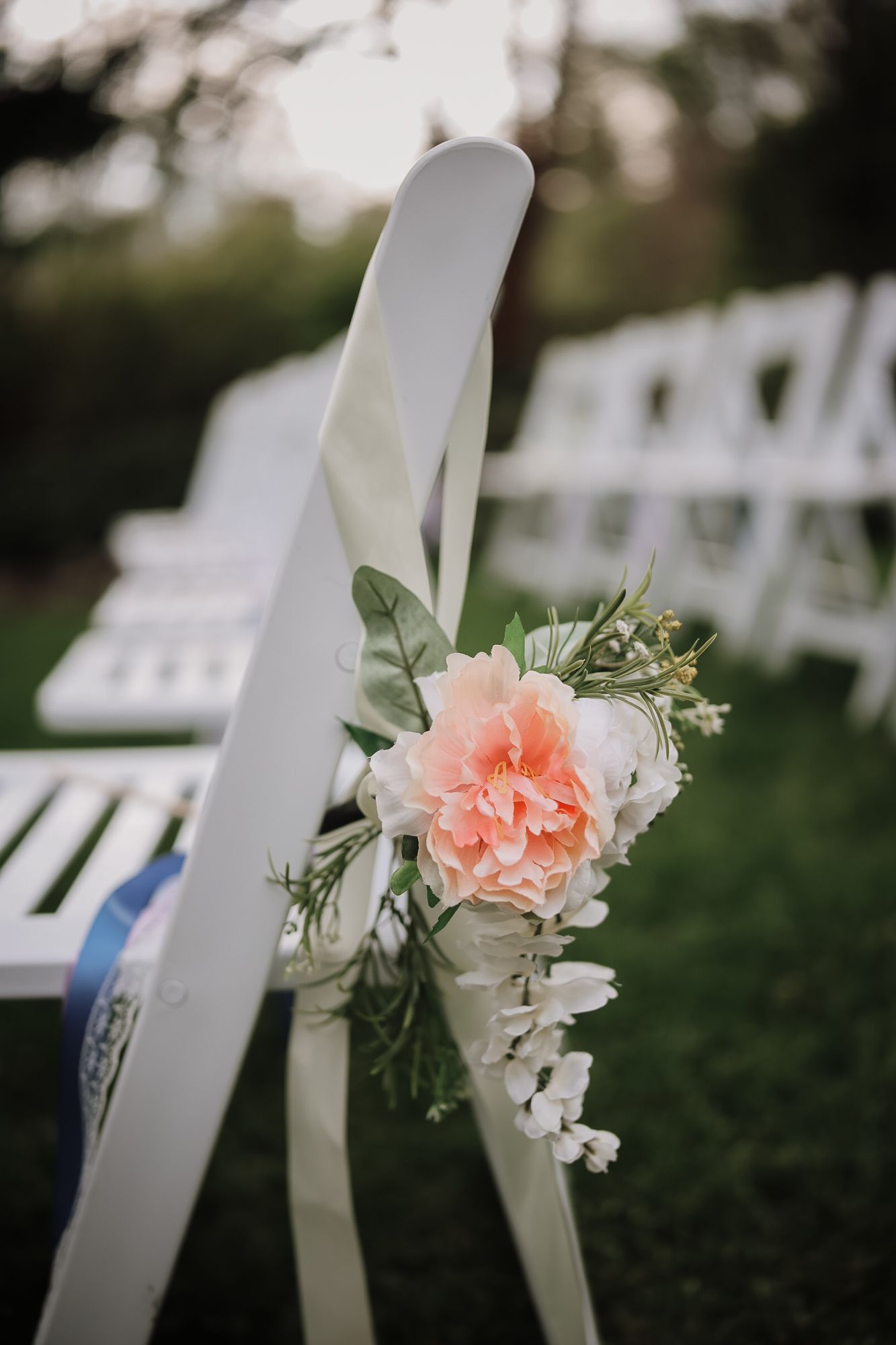 White folding chairs adorned with peach roses and white floral arrangements line an outdoor wedding ceremony aisle.