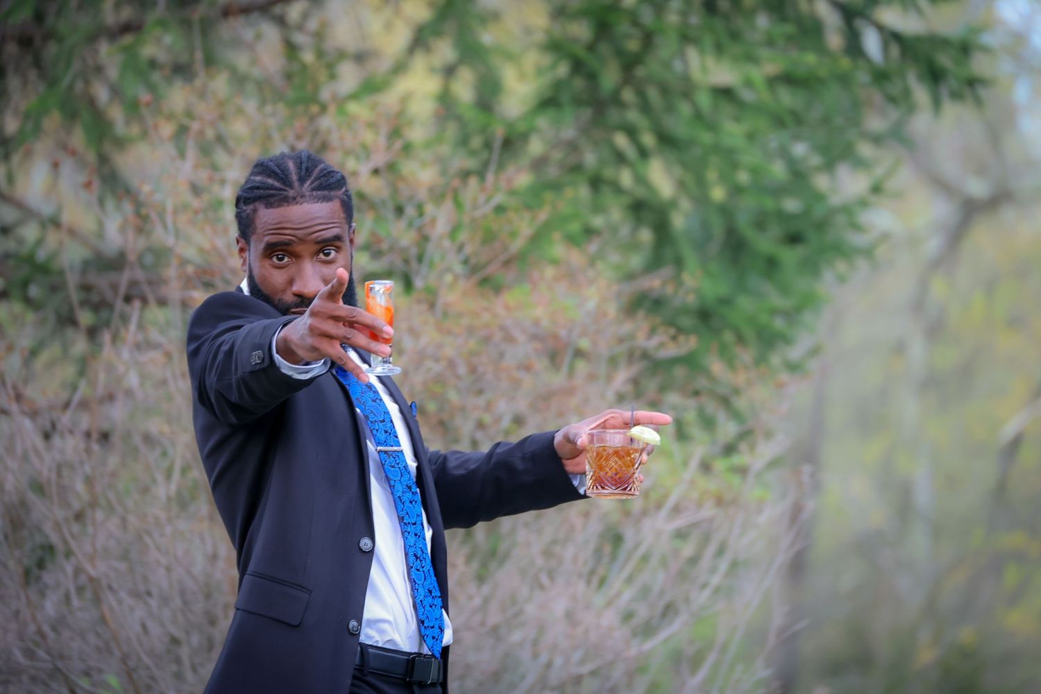 Person in black suit pointing finger gun while holding a drink outdoors near green trees.