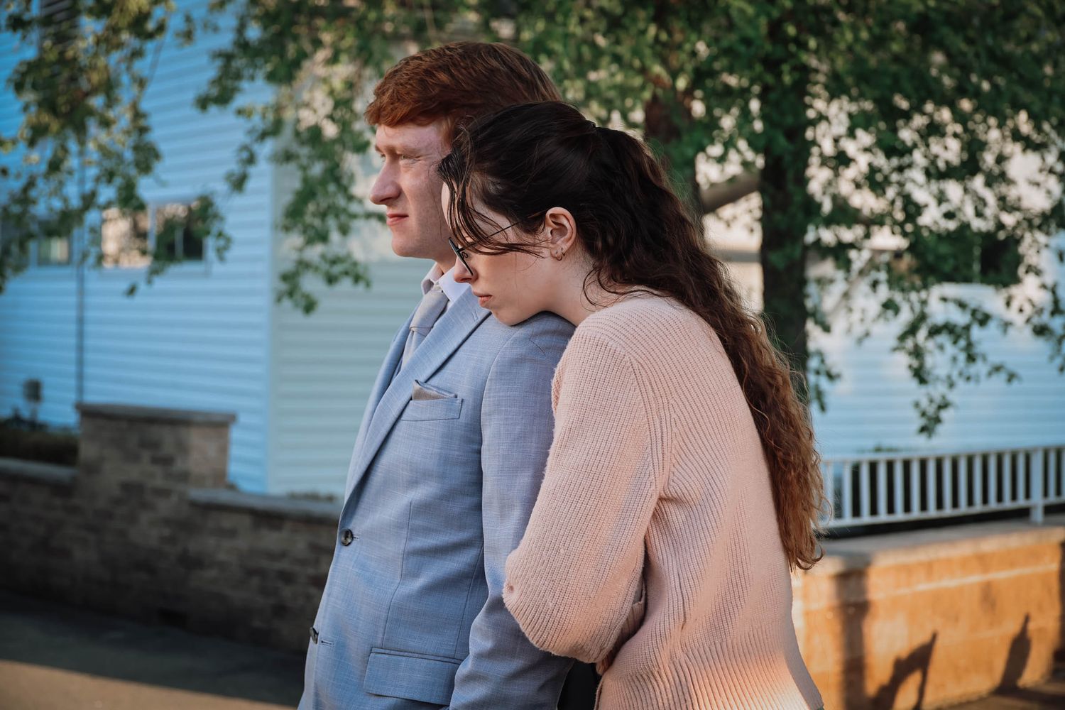 A young couple in formal attire share a tender moment outdoors near a blue building at sunset.