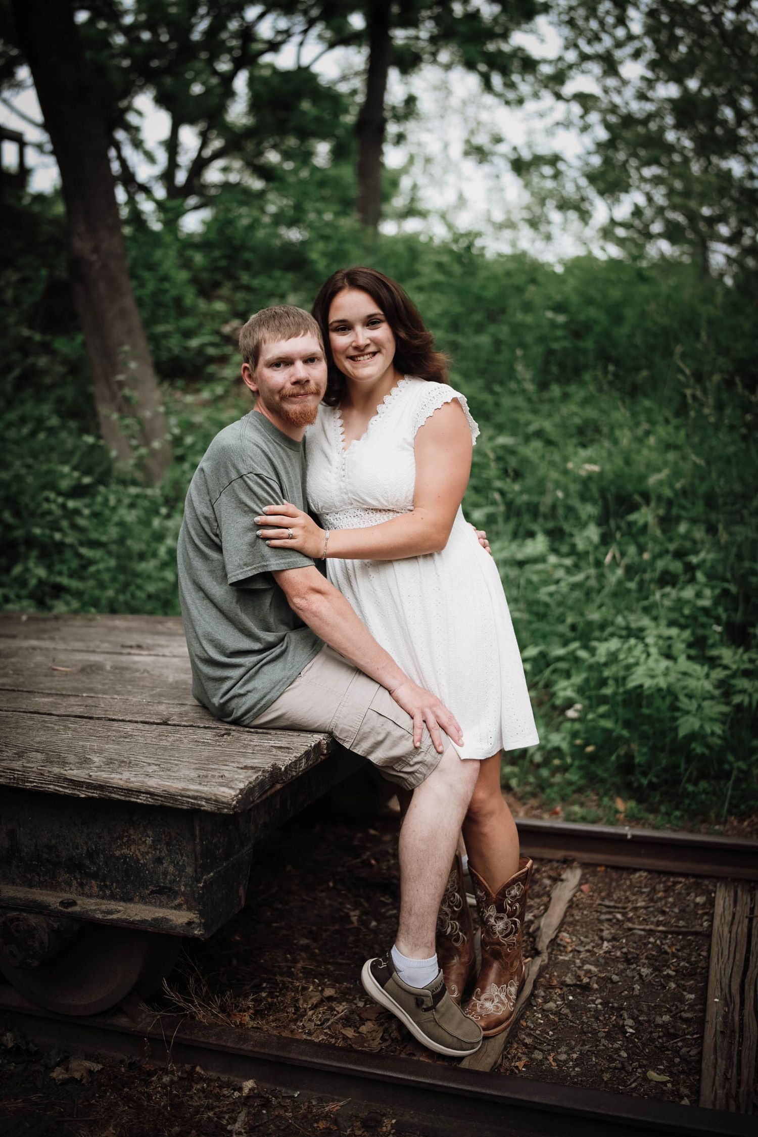 Couple sitting on railroad tracks surrounded by lush greenery, woman in white dress and cowboy boots.
