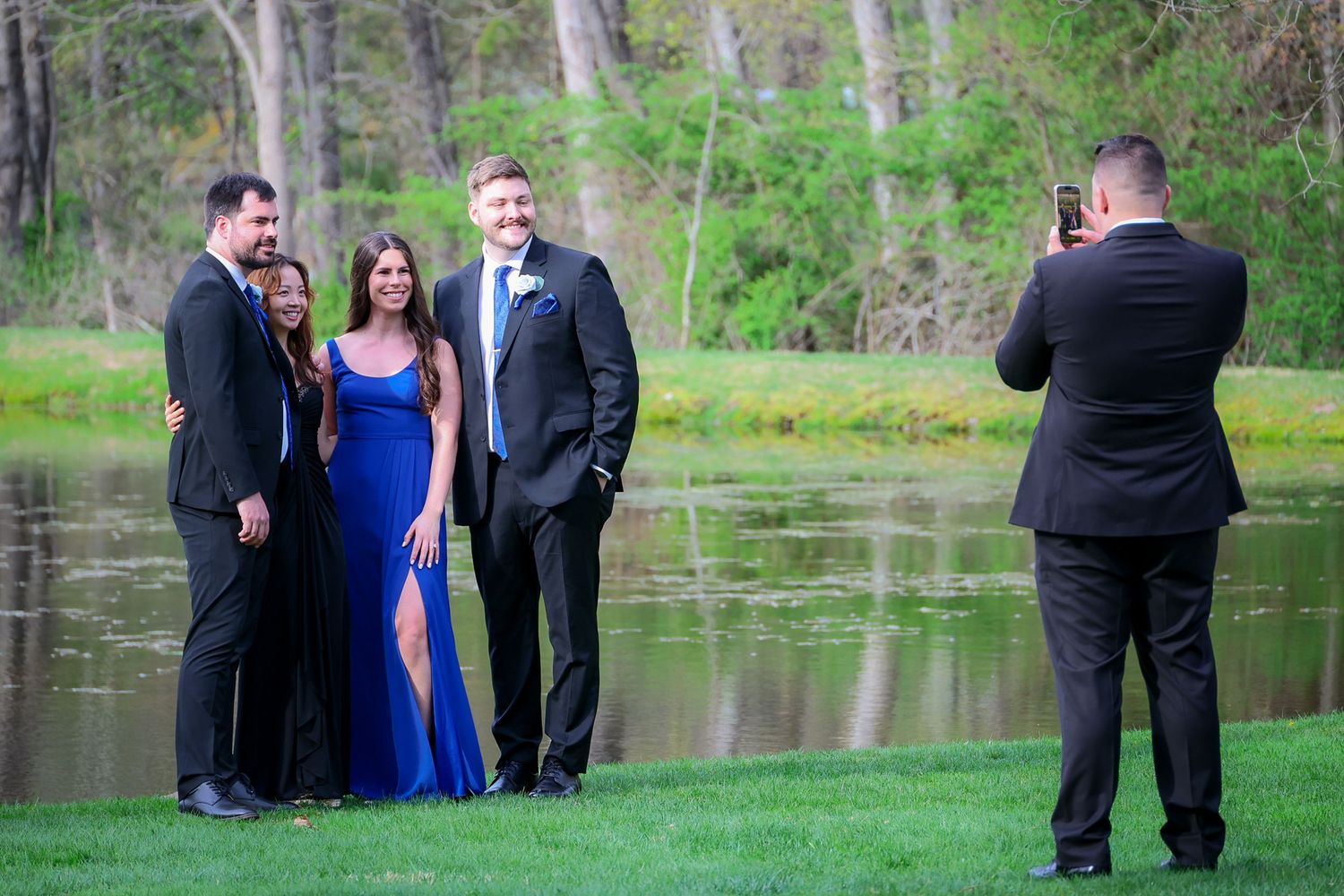 Formally dressed group posing for photos by a pond, with a man in blue dress photographed by photographer outdoors.