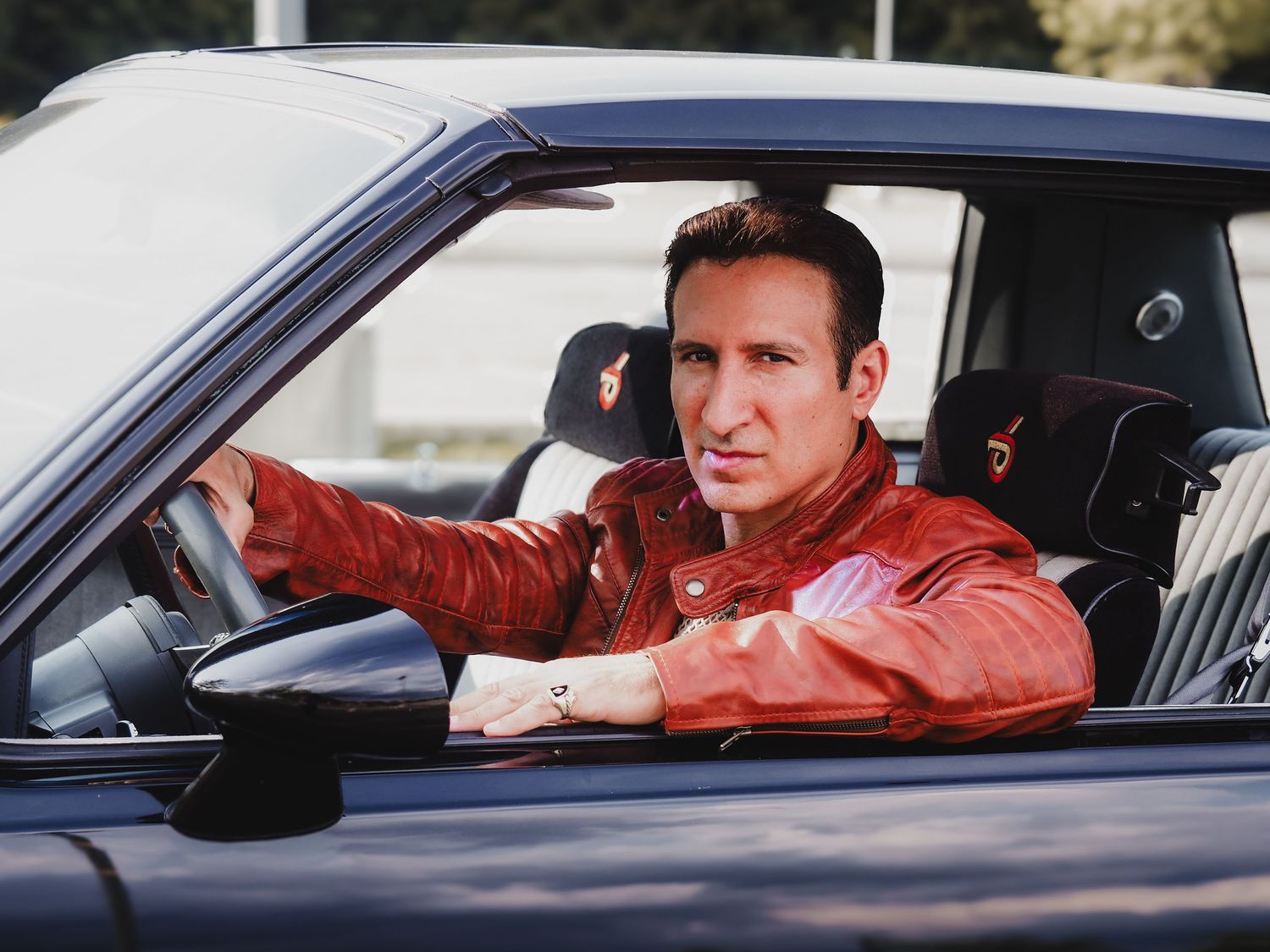 Driver wearing a red leather jacket sits behind the wheel of a black sports car.
