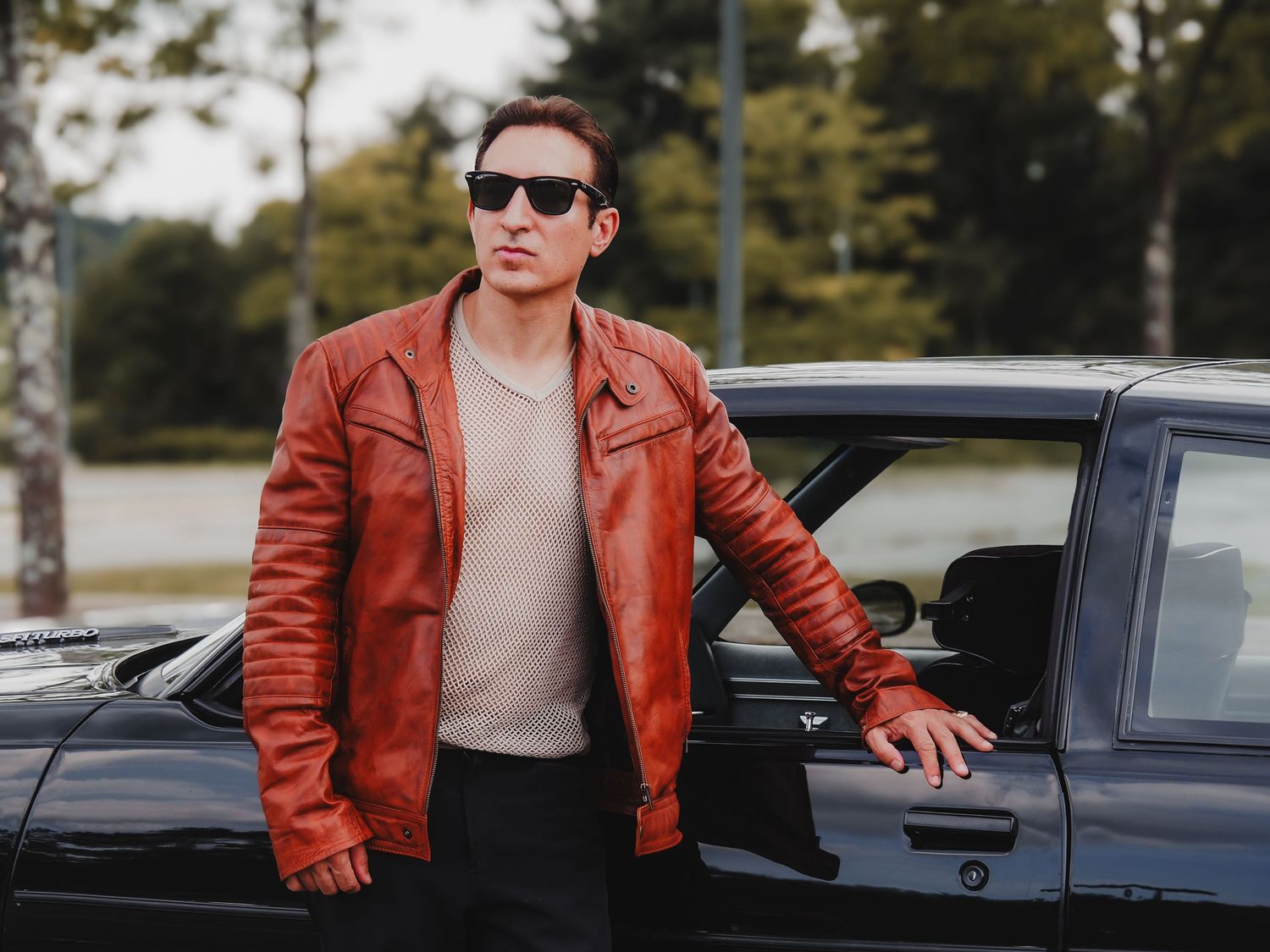 A person in a brown leather jacket and sunglasses poses next to a parked car in an outdoor setting.