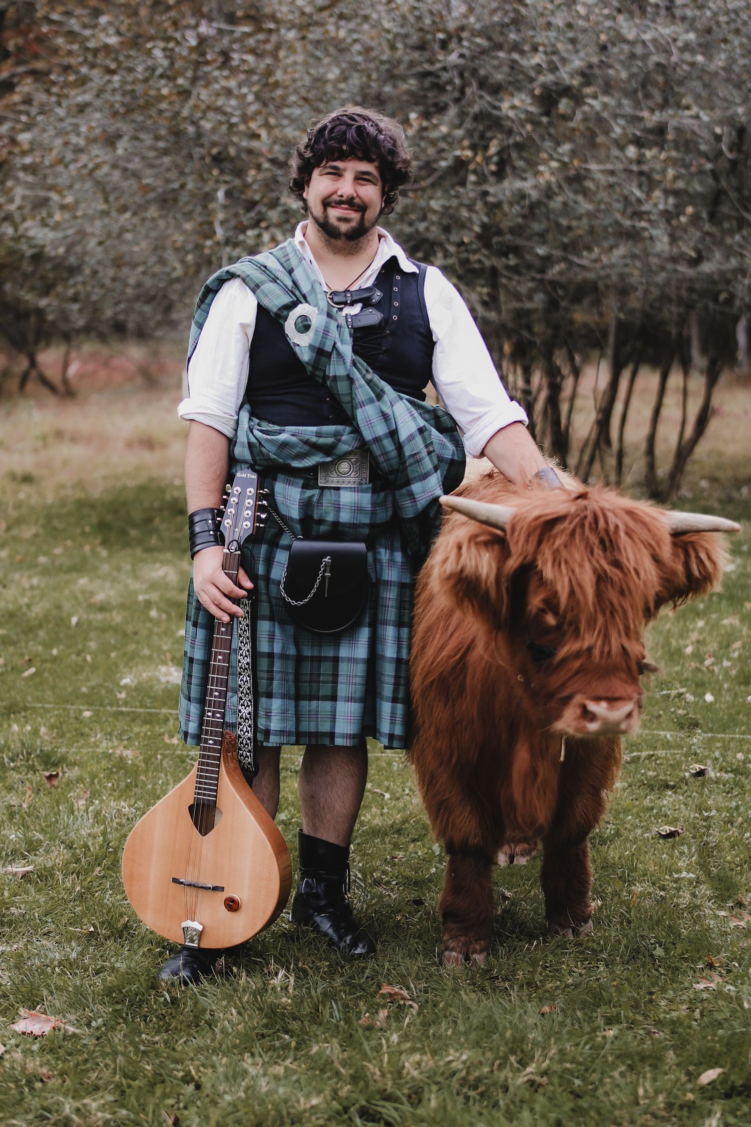 Traditional Scottish musician in kilt and vest poses with acoustic instrument next to Highland cow in grassy field.
